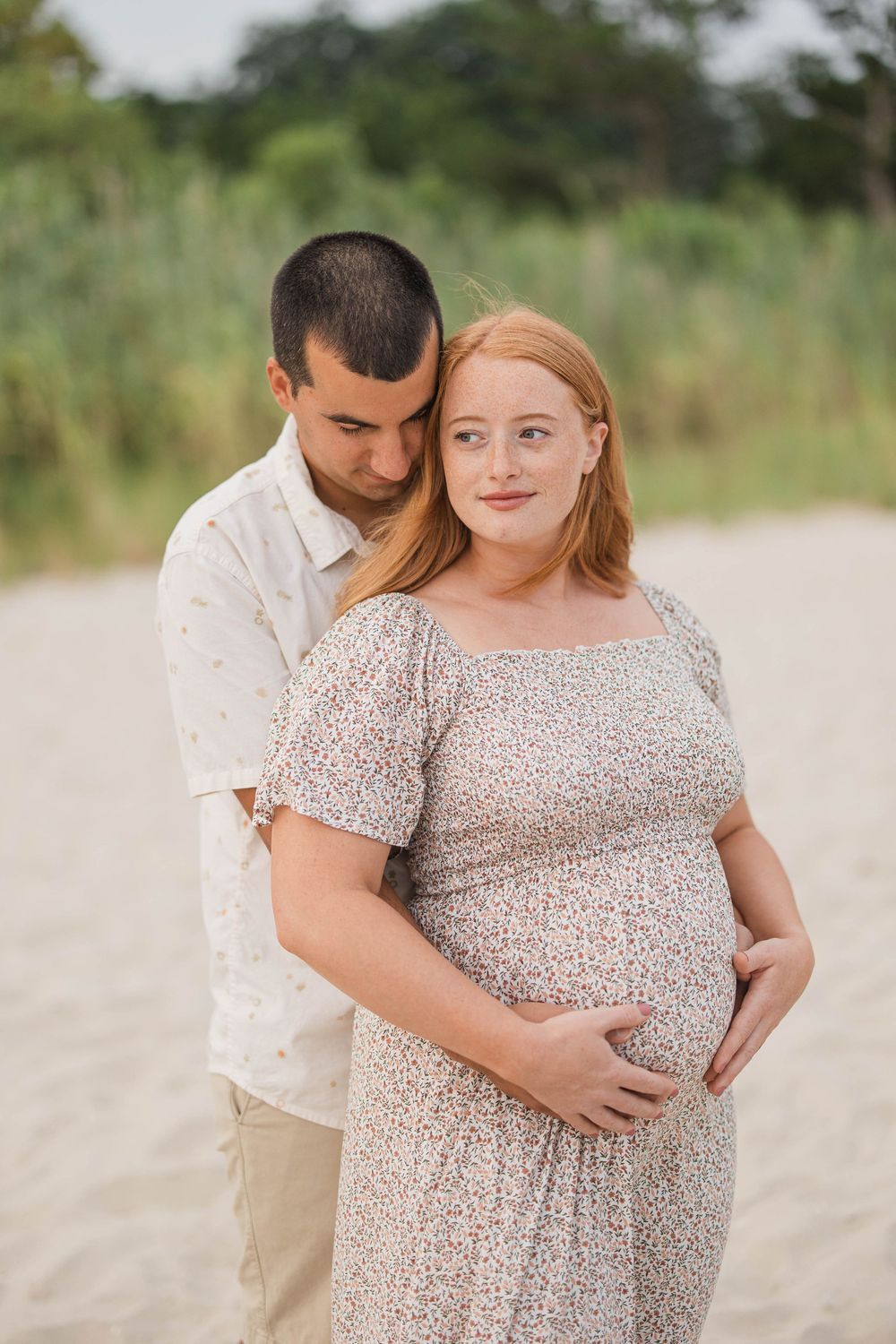 Pregnant couple embracing on a sandy beach, woman in floral dress cradling her bump, man resting his head on her shoulder.