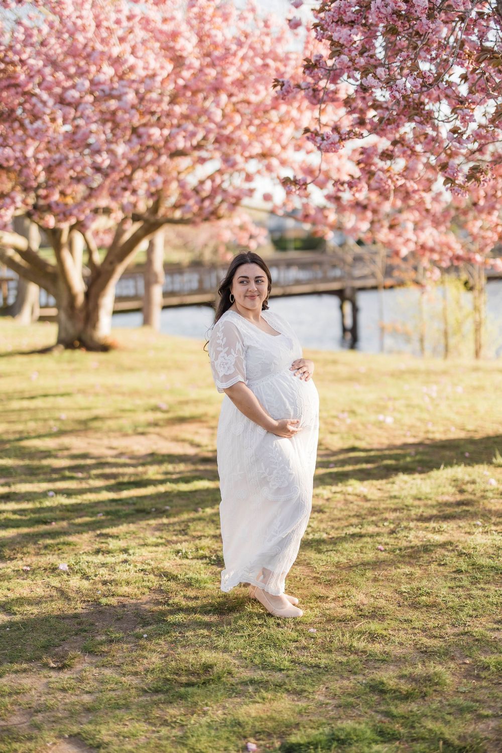 Pregnant woman in white dress cradling baby bump under pink cherry blossom trees in a sunny park.