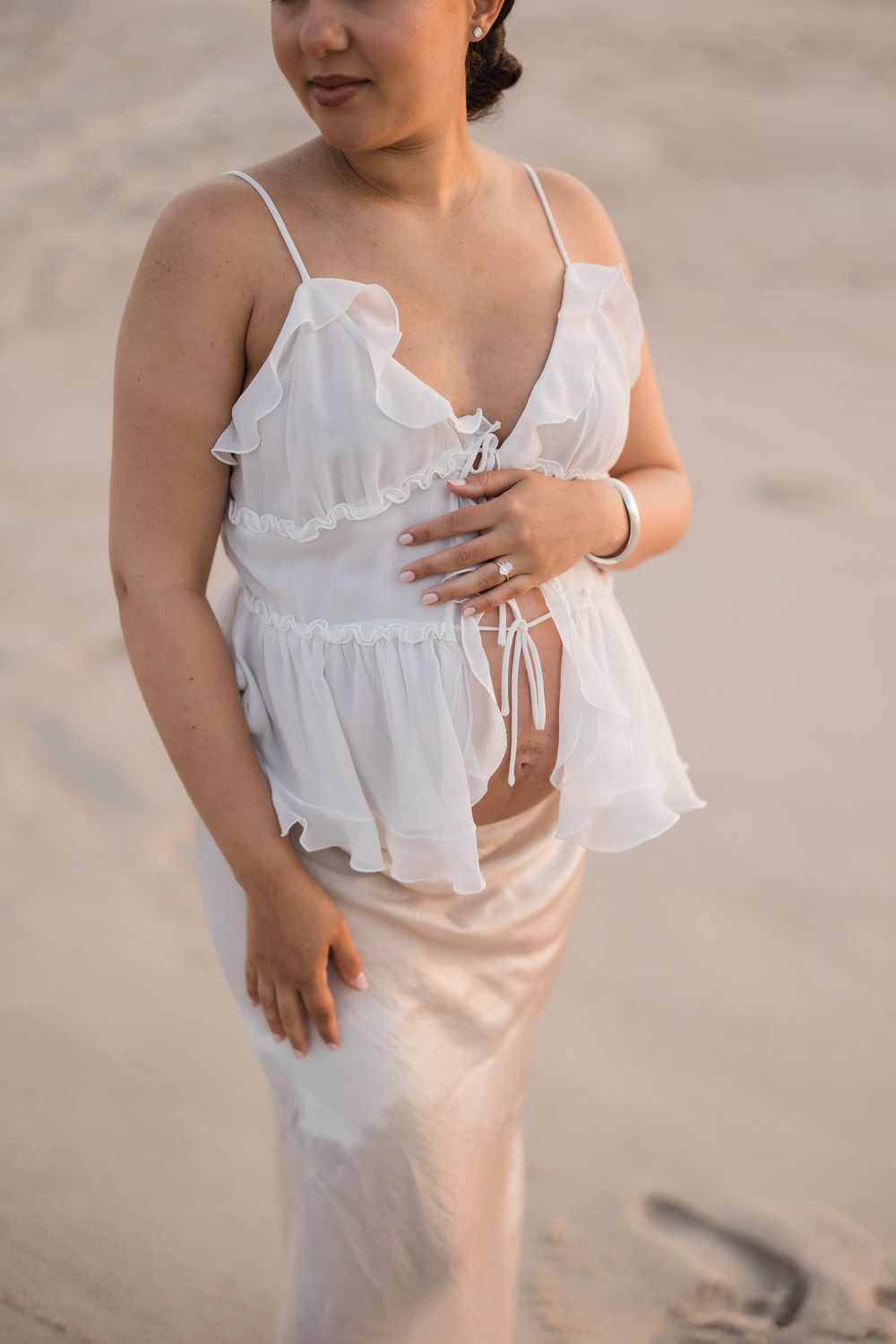 Woman in white sheer ruffled top and satin skirt standing on sandy beach in soft golden light.