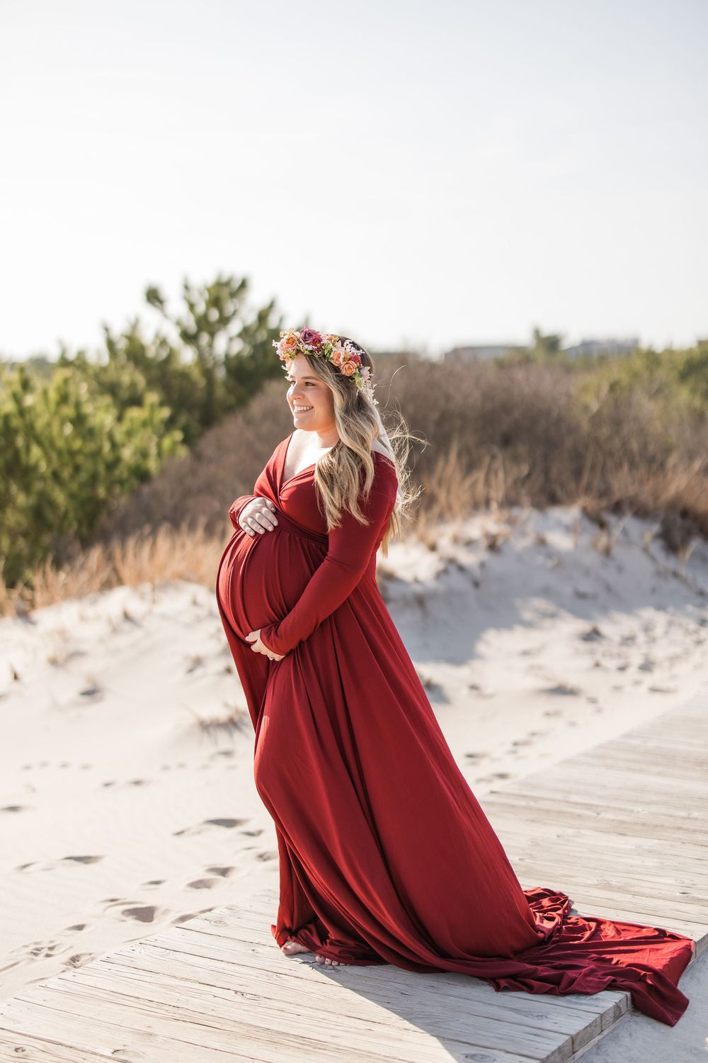 Pregnant woman in red gown and floral crown poses on sandy beach boardwalk during golden hour maternity photo session.
