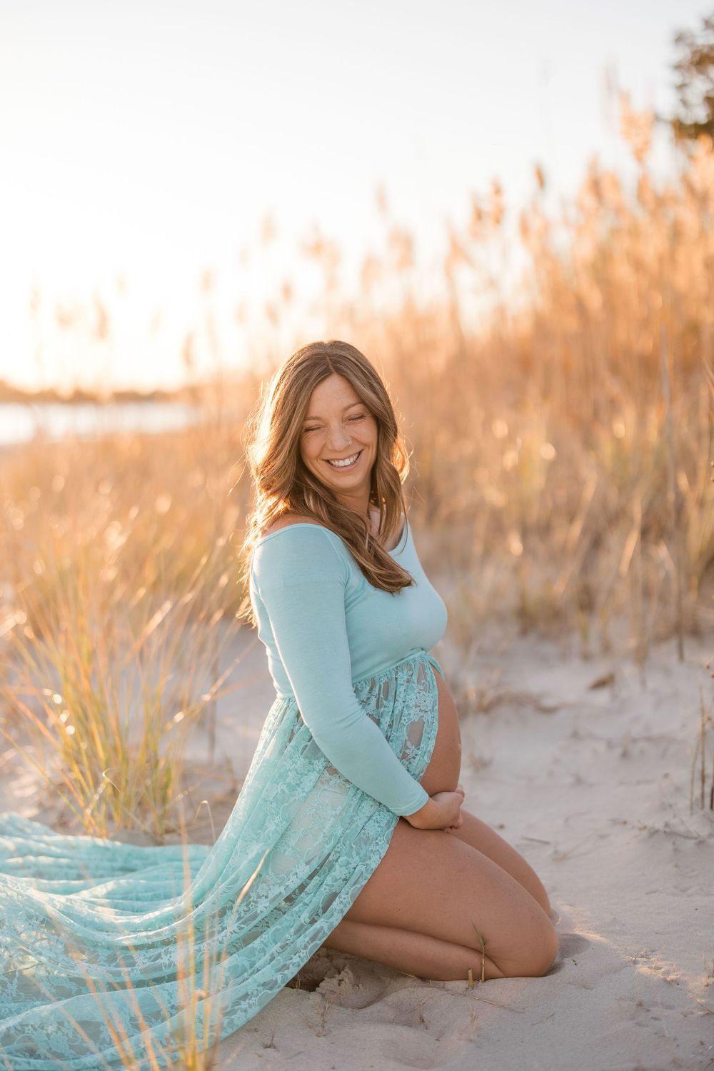Joyful pregnant woman in mint lace dress kneeling on sandy beach dunes at sunset golden hour.
