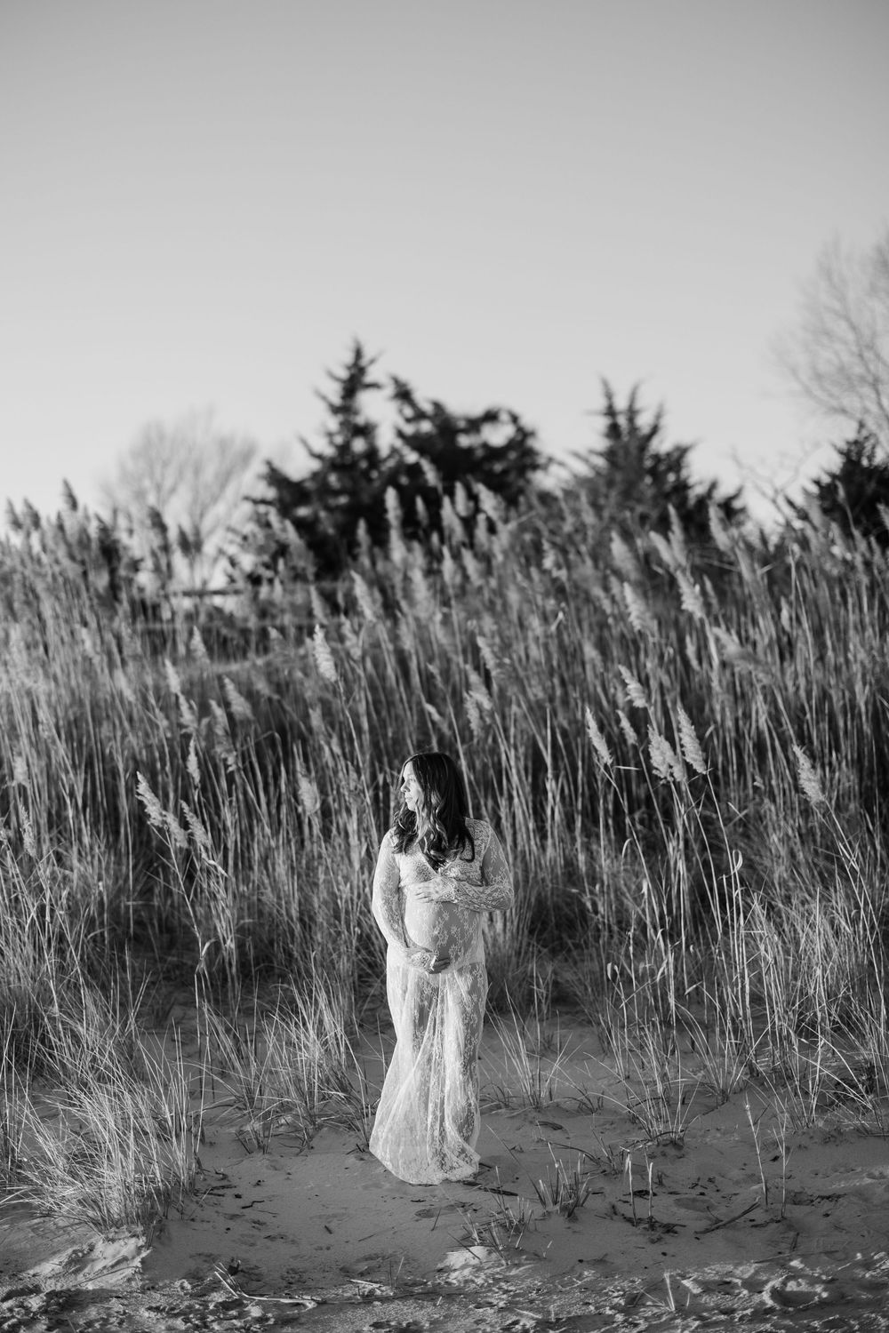 Black and white photo of a pregnant woman in a lace gown standing among tall reeds on a sandy shore.