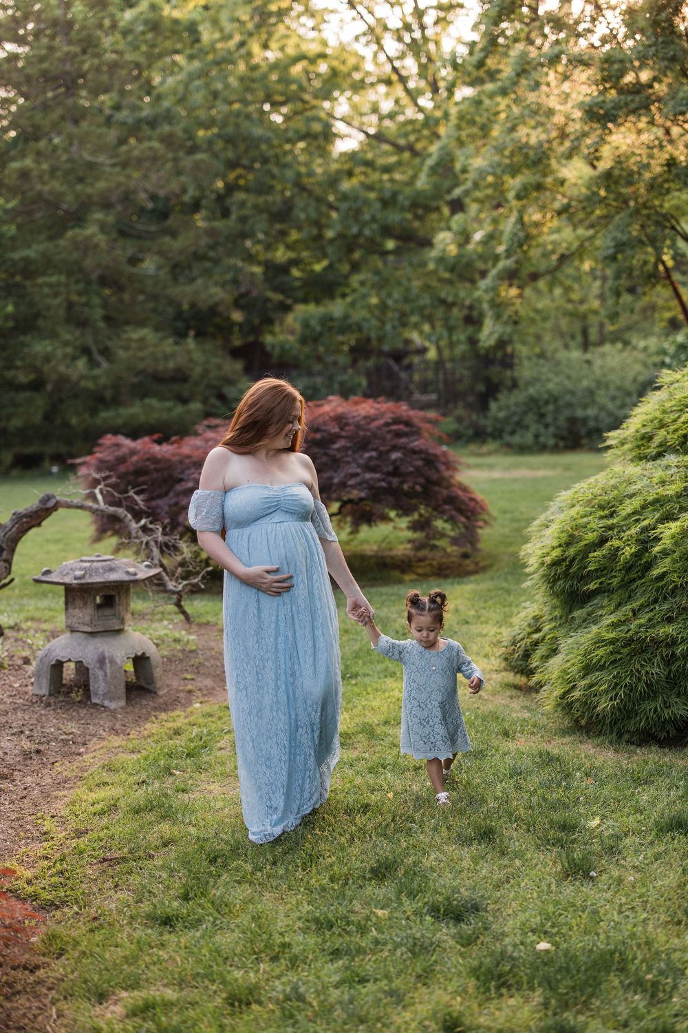 Pregnant woman in blue dress holds hands with toddler girl in garden with Japanese maple trees.