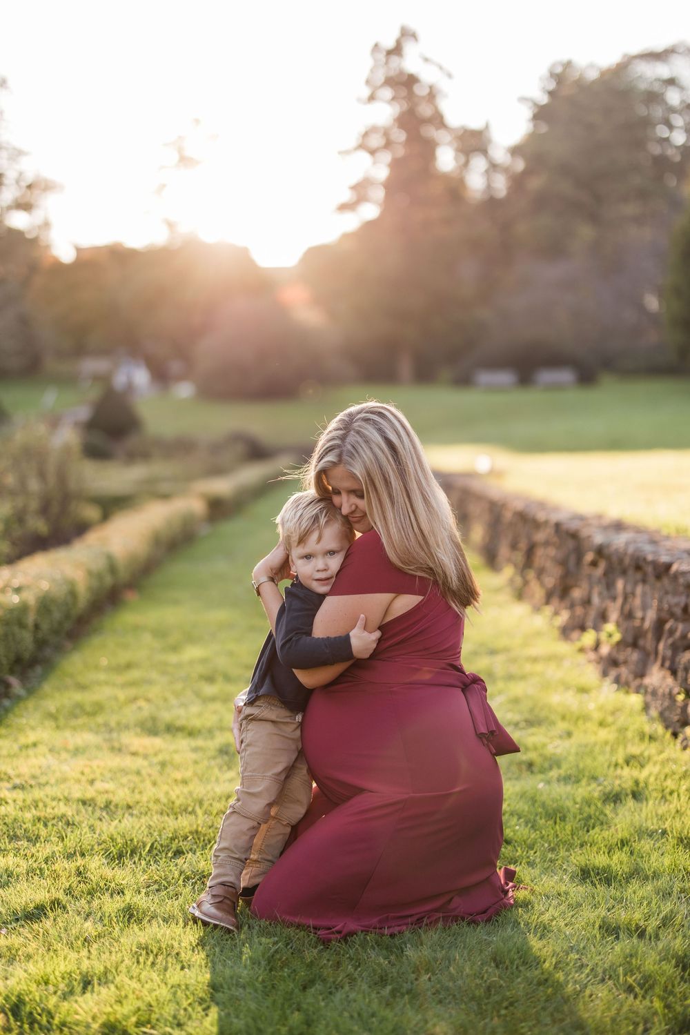 A mother in a red dress hugs her toddler son on a garden lawn during golden hour sunset.