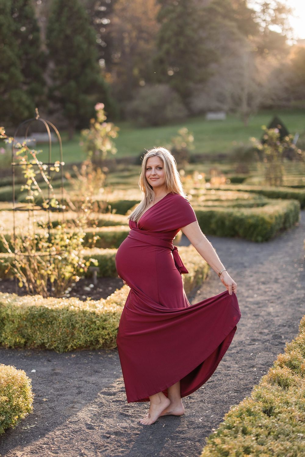 Elegant pregnant woman in flowing burgundy wrap dress twirling barefoot in a formal garden at sunset.