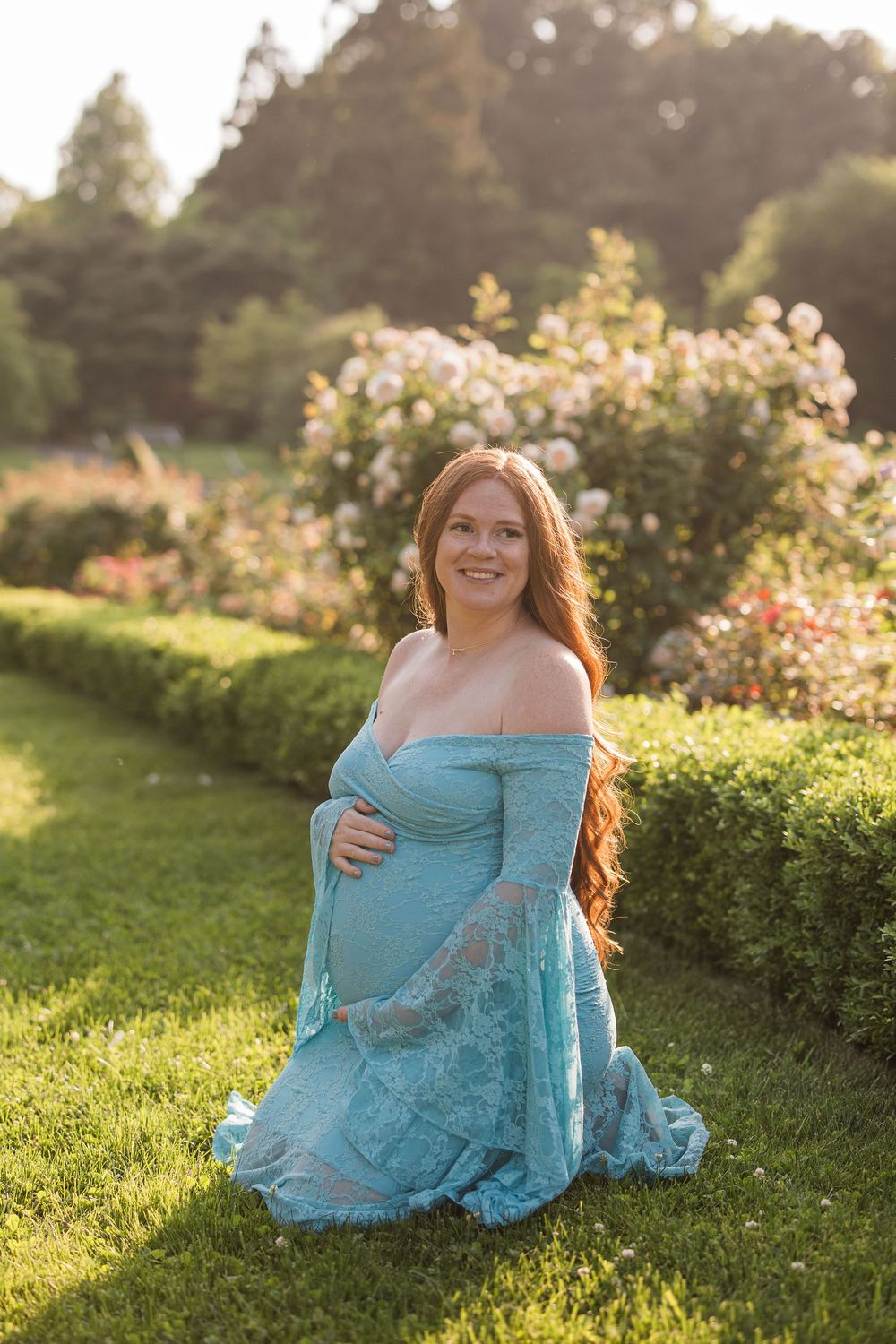 Smiling pregnant woman in teal lace gown kneeling in garden with blooming roses and hedges in golden hour light.