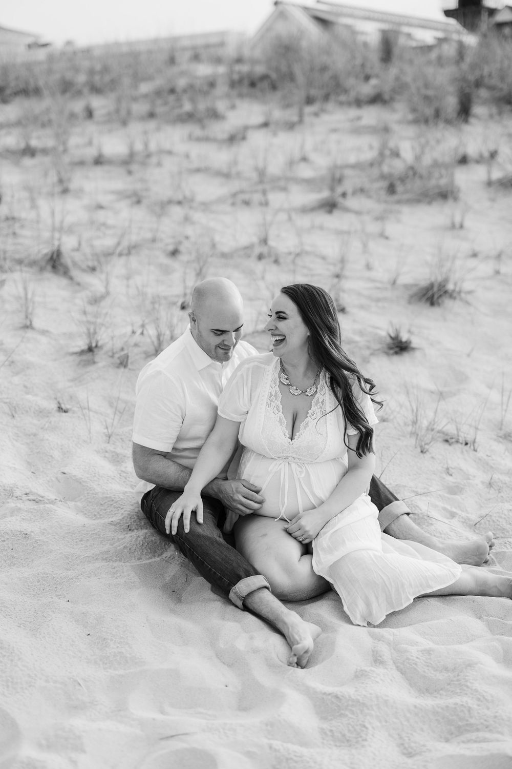 Black and white photo of expectant couple sitting together on sandy beach smiling at each other.