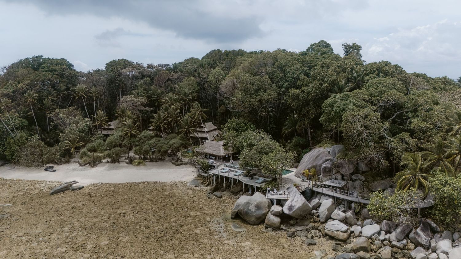 Aerial view of a tropical resort nestled among lush jungle trees with wooden structures along a rocky beach.