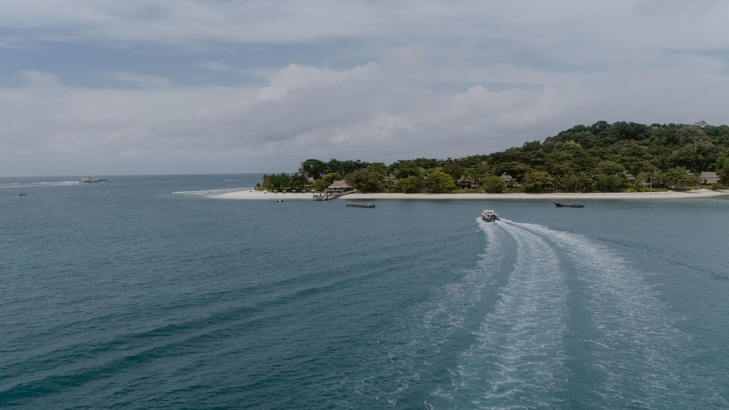 Aerial view of a speedboat leaving a wake near a lush tropical island under a cloudy sky.