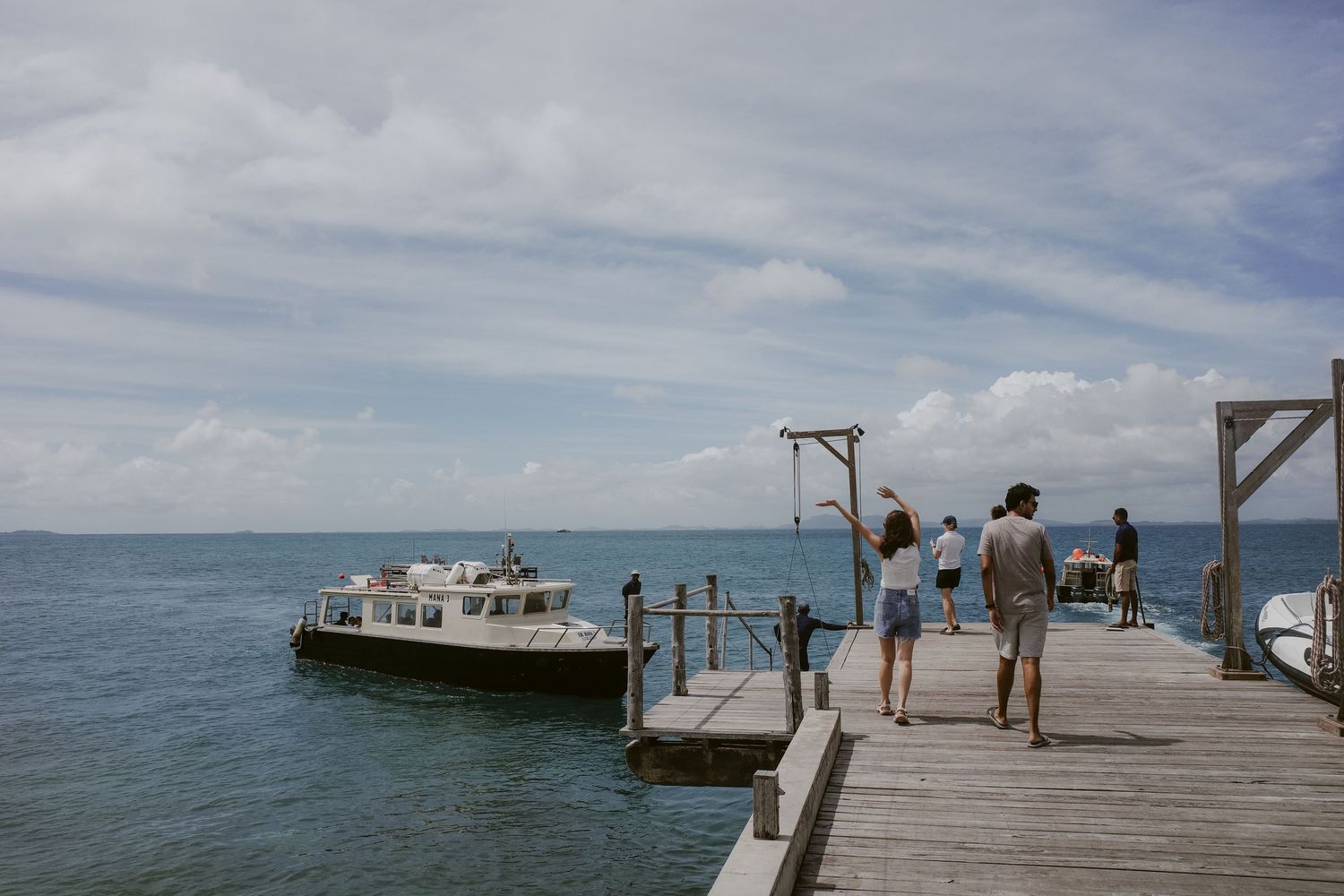 Group of people standing on a wooden dock watching a white boat arrive on calm turquoise waters.