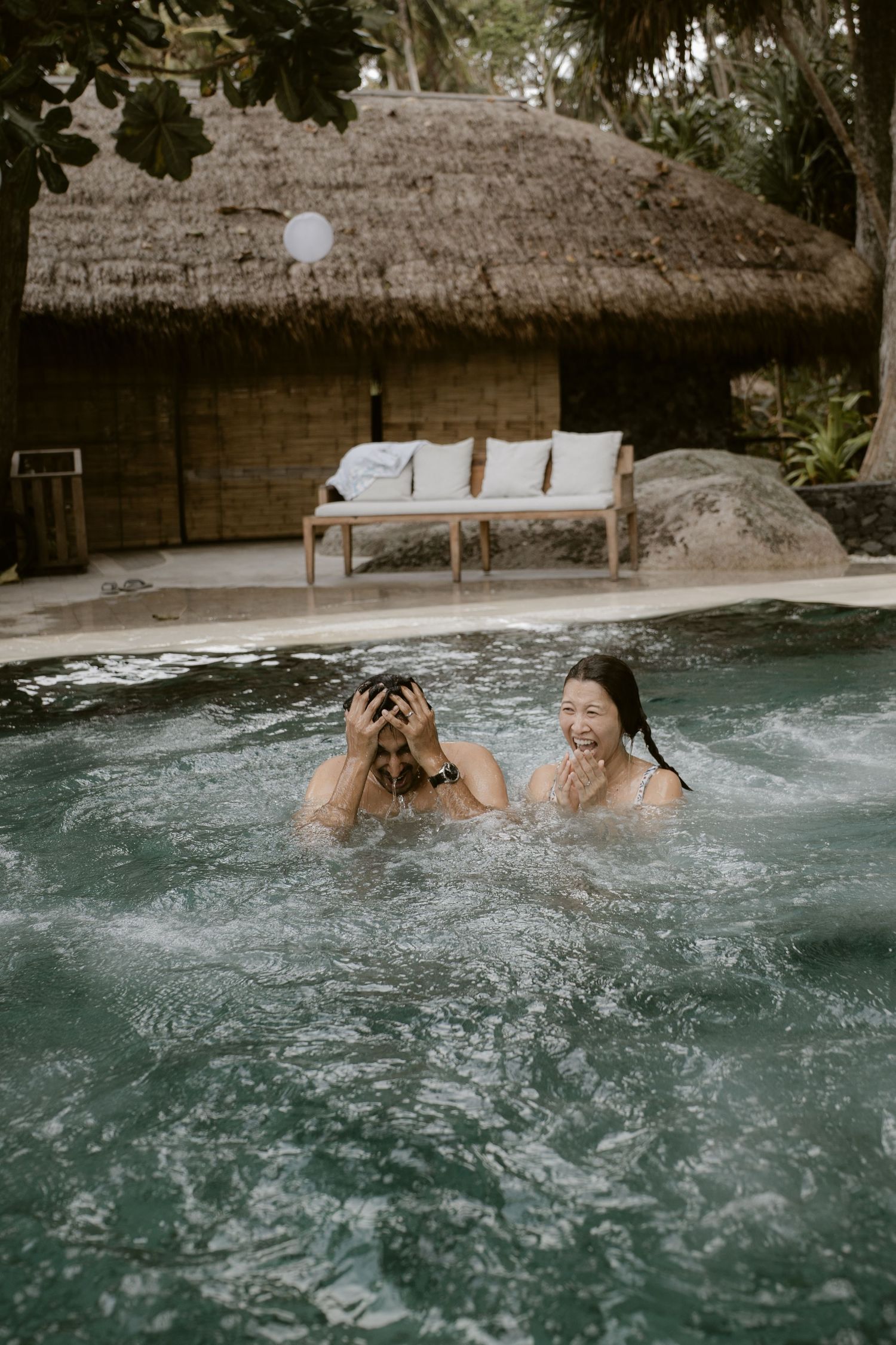 Two people laughing and splashing in a tropical resort pool with a thatched roof bungalow behind them.
