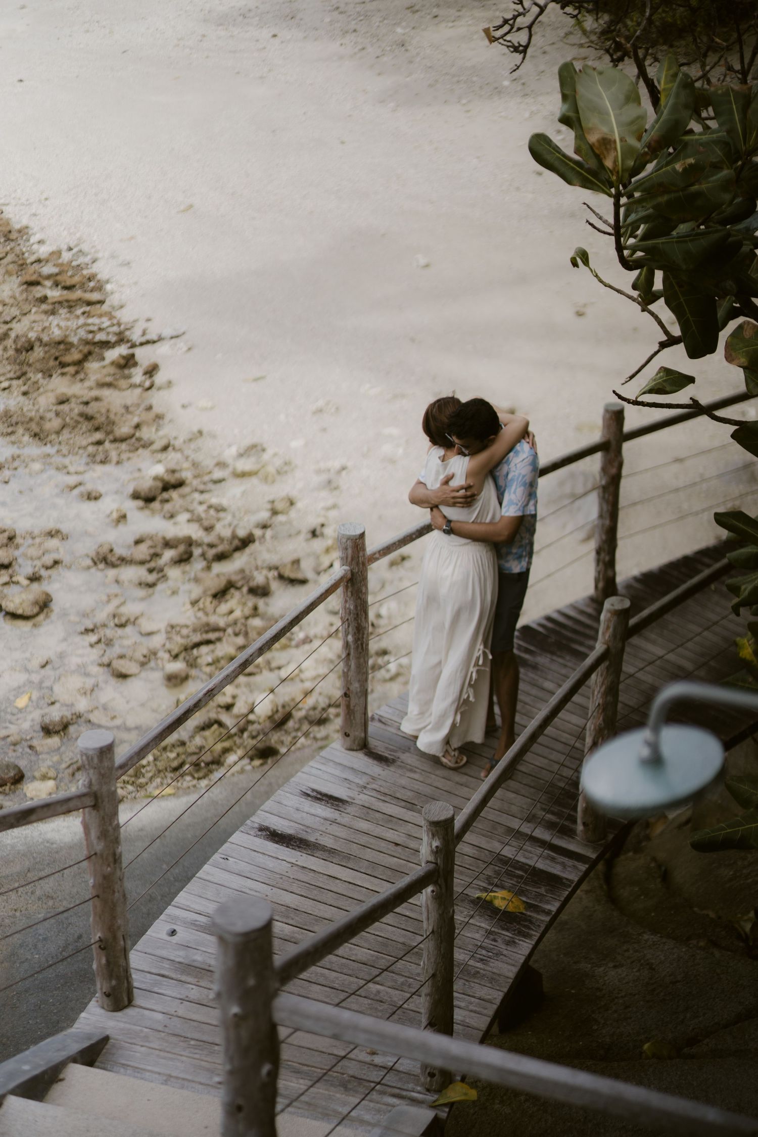 Bride and groom share tender embrace on wooden boardwalk overlooking sandy tropical beach.