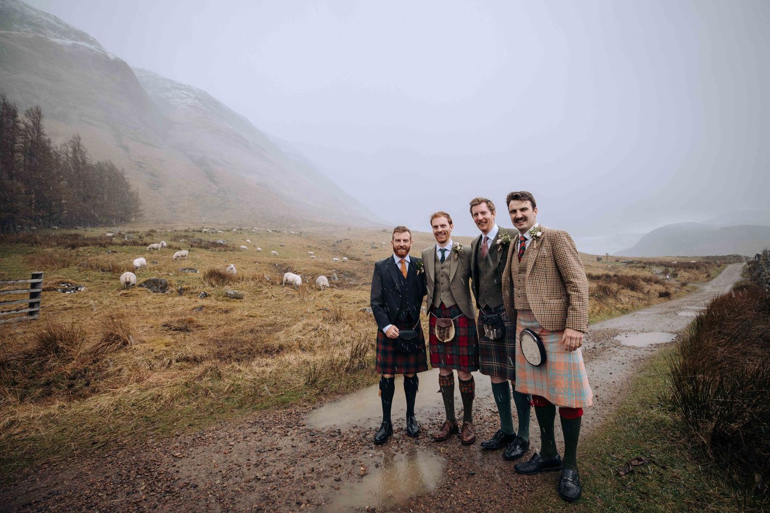 Four men wearing traditional Scottish kilts posing together on a misty Highland path with mountains in the background.