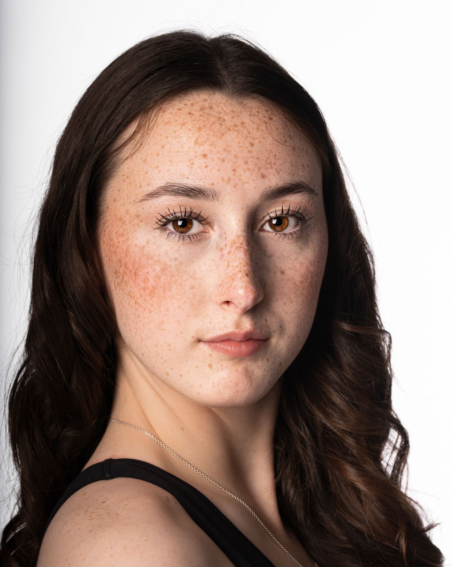 Young woman with freckles, hazel eyes, and dark wavy hair poses for a portrait against a white background.