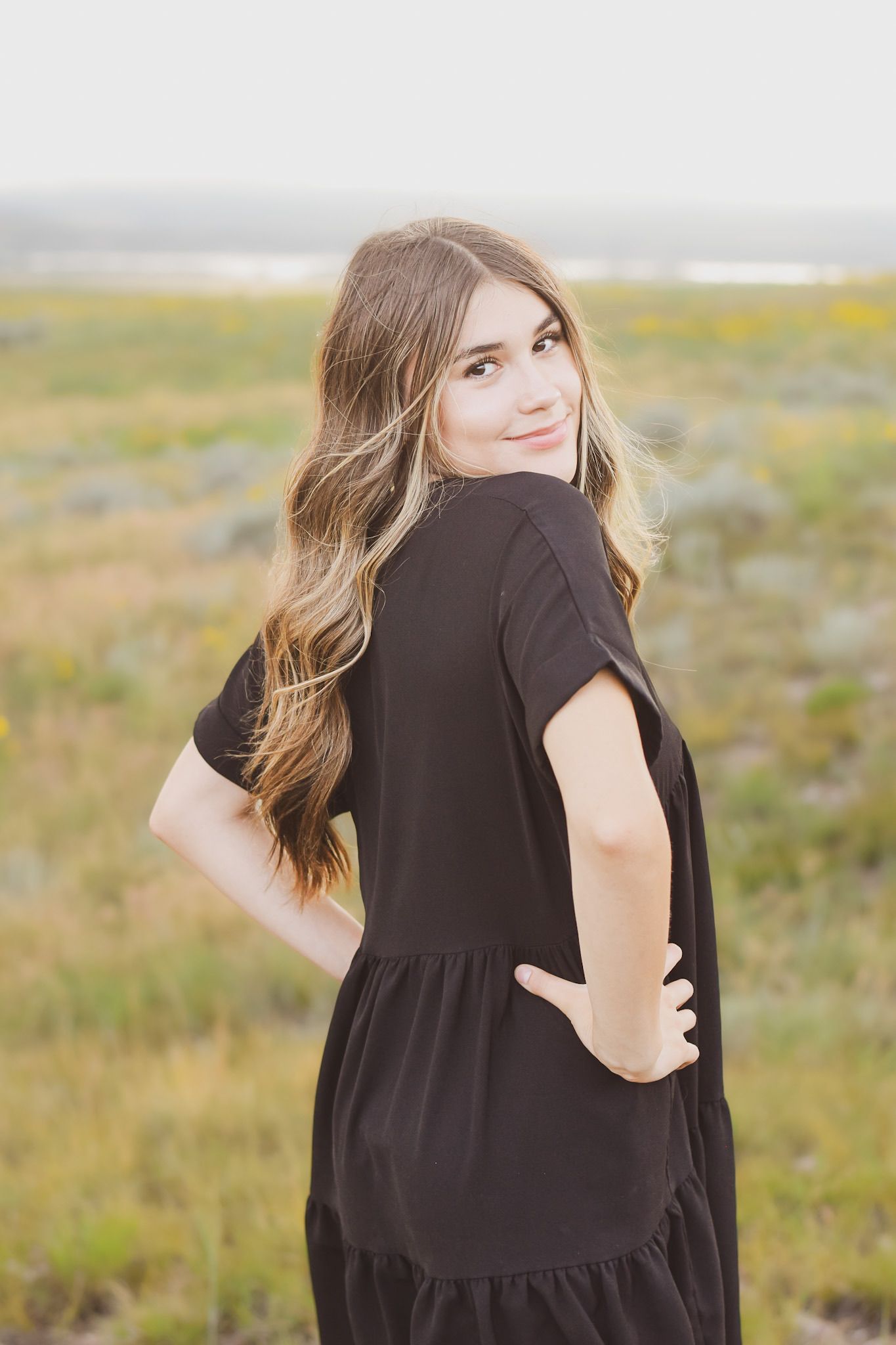 Young woman in black dress poses in a wildflower field, looking back over her shoulder with a smile.