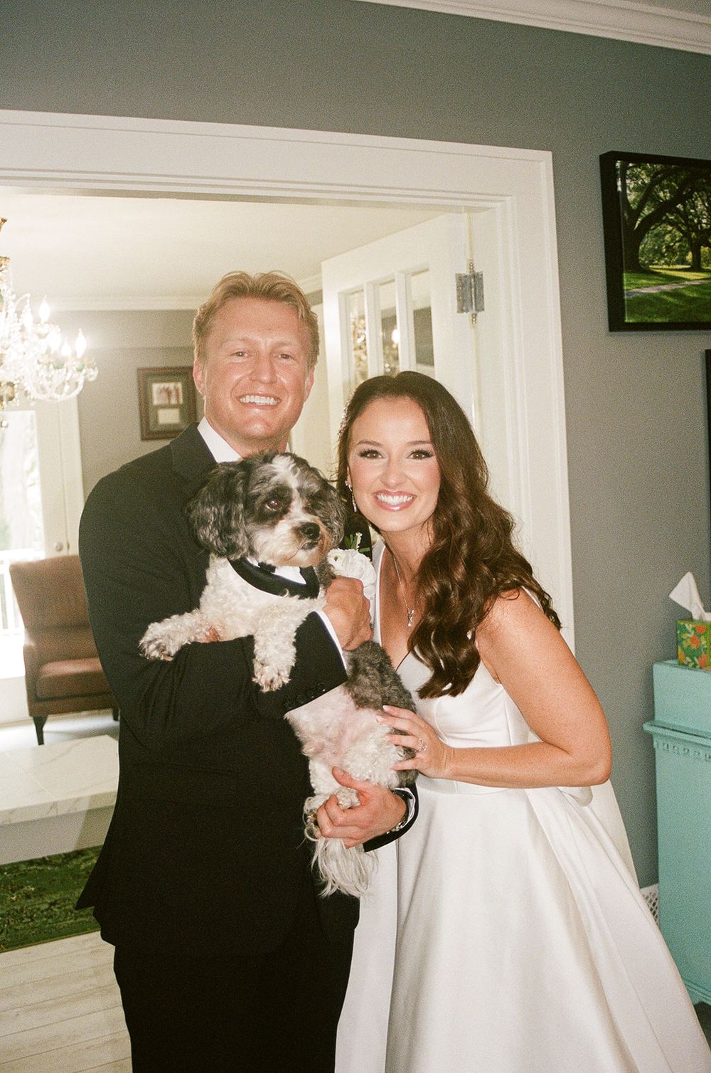 Bride and groom in wedding attire smiling while holding their small dog dressed in a tuxedo inside a home.