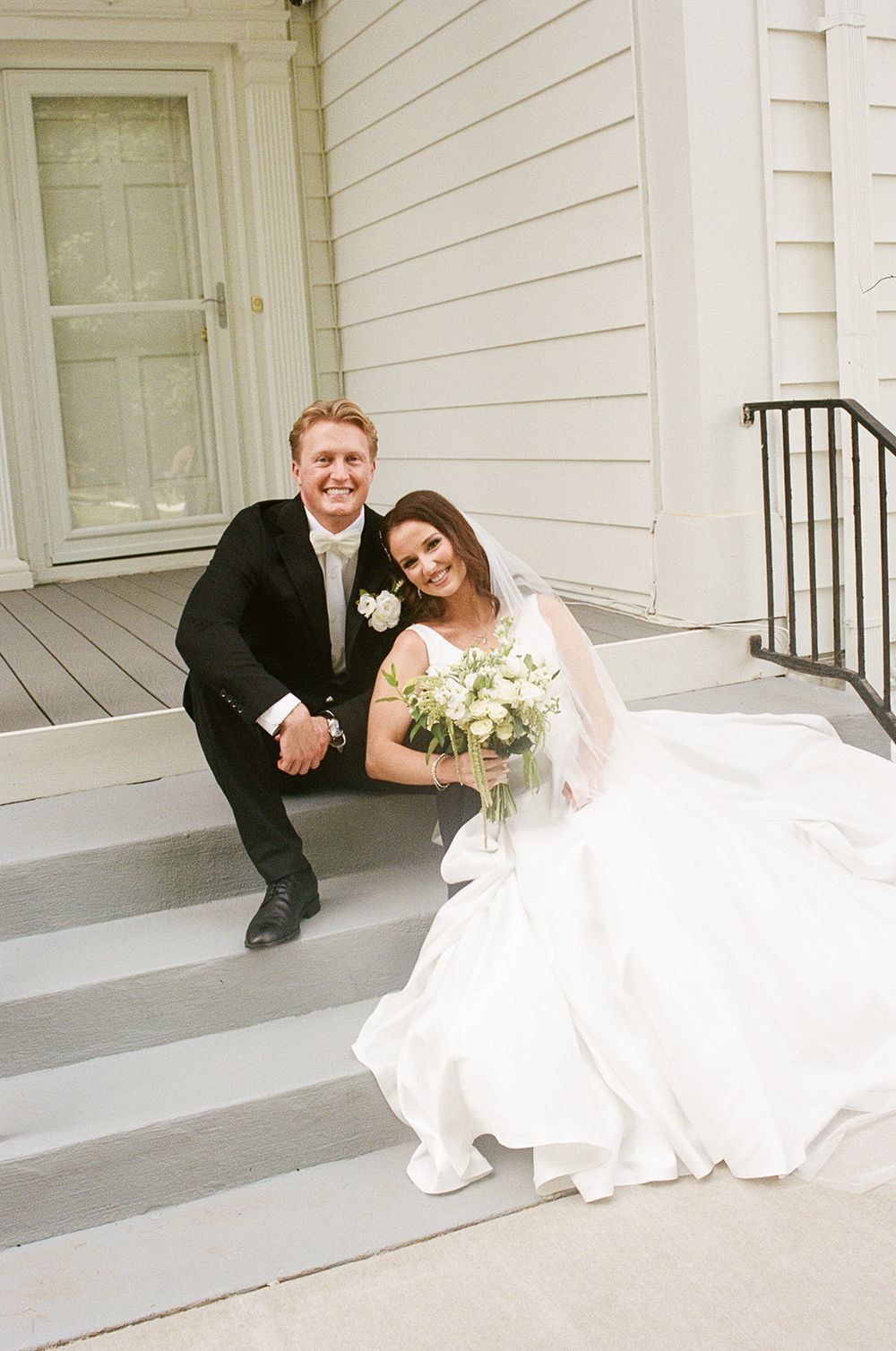 Bride and groom sitting on white steps, she holds white bouquet in wedding dress, he wears black suit with boutonniere.
