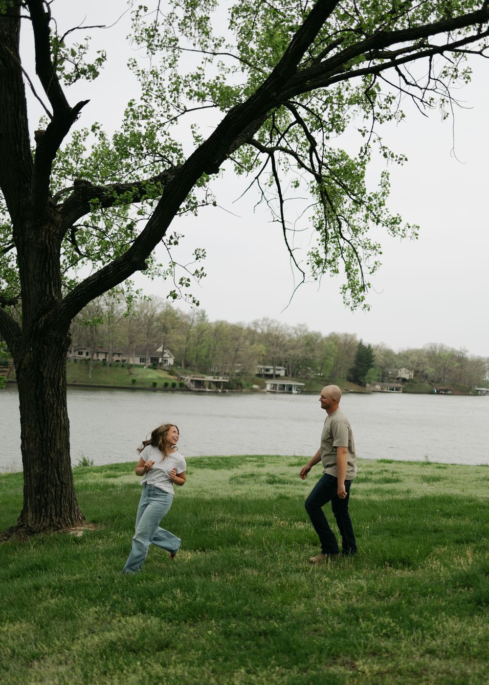 Couple playfully running toward each other on green grass near a lakeside tree during engagement photos.