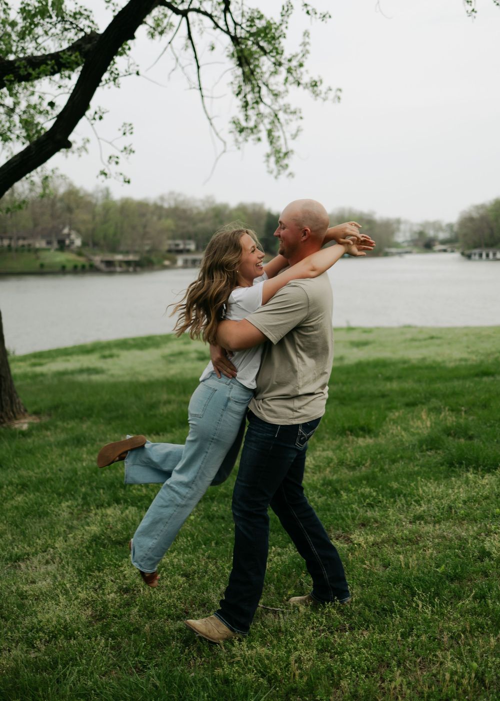 Man lifting and spinning woman in a joyful embrace near a lake during an outdoor engagement session.