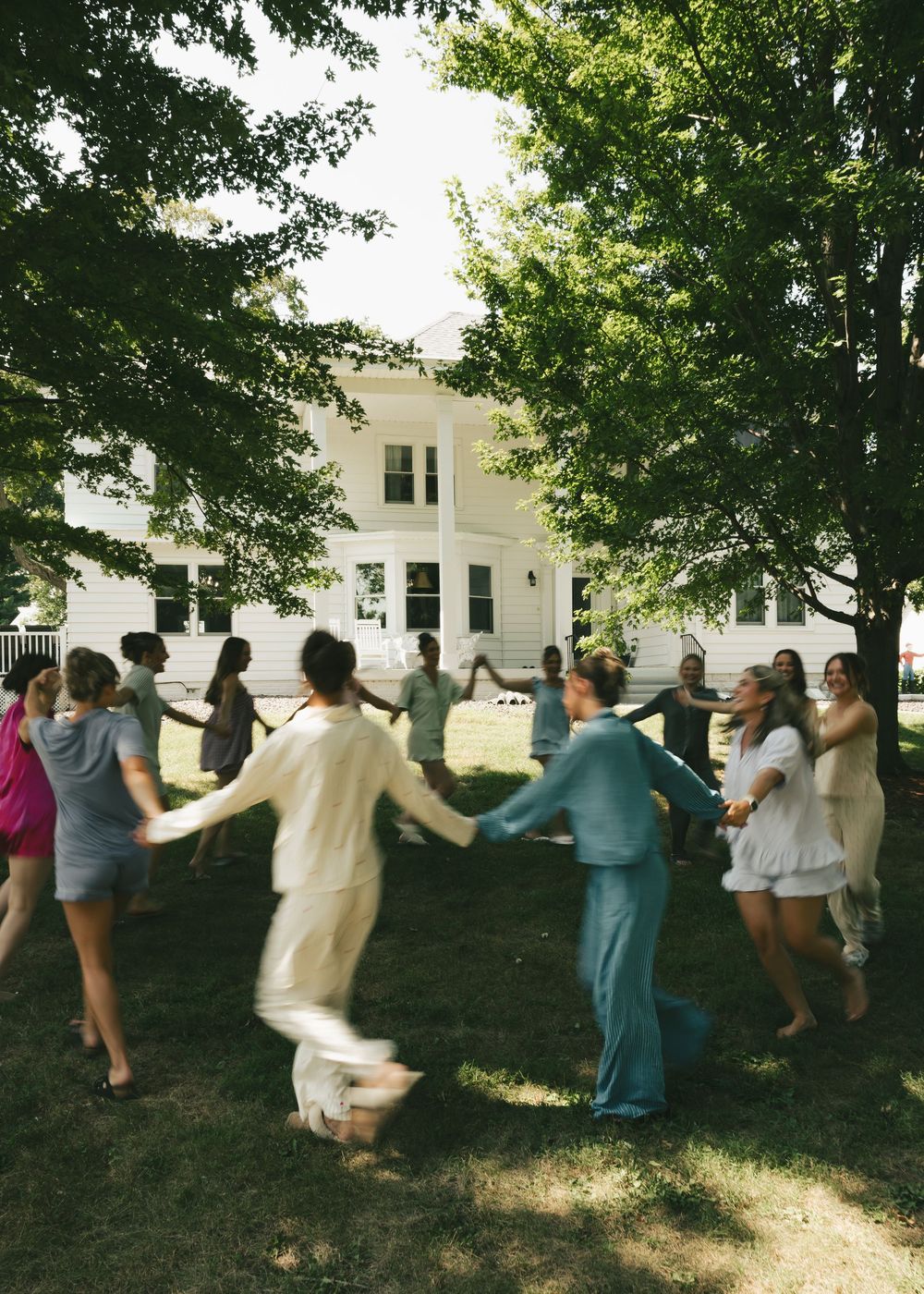 Group of wedding guests holding hands in a circle dancing outdoors under trees near a white farmhouse.