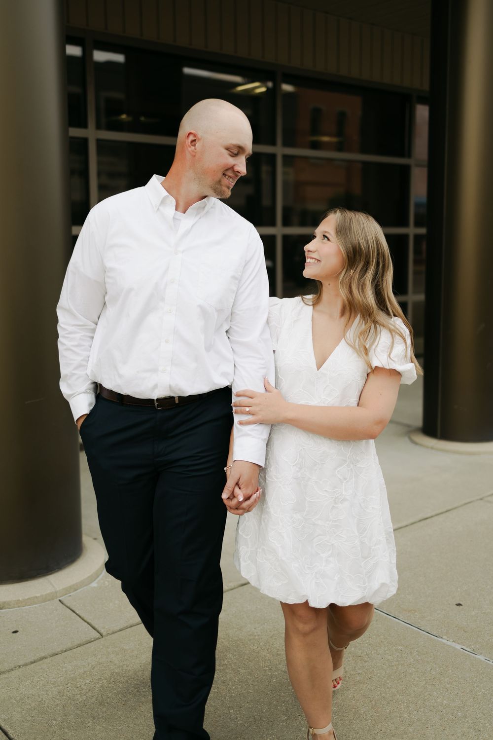 Couple in matching white outfits holding hands and smiling at each other while walking outside a modern building.