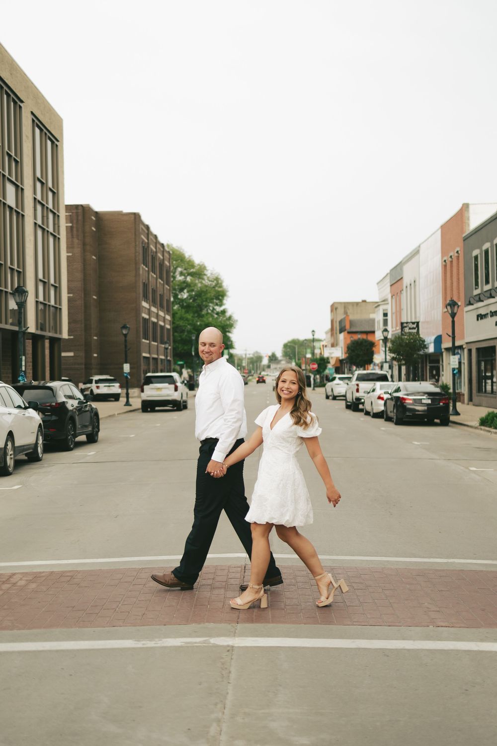 Happy couple in white outfits holding hands while crossing a charming small-town street during engagement photos.