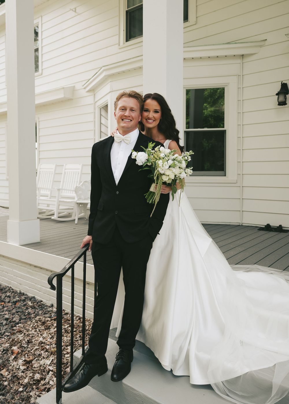 Bride and groom posing on white farmhouse porch steps, bride in elegant white gown with train holding white bouquet.
