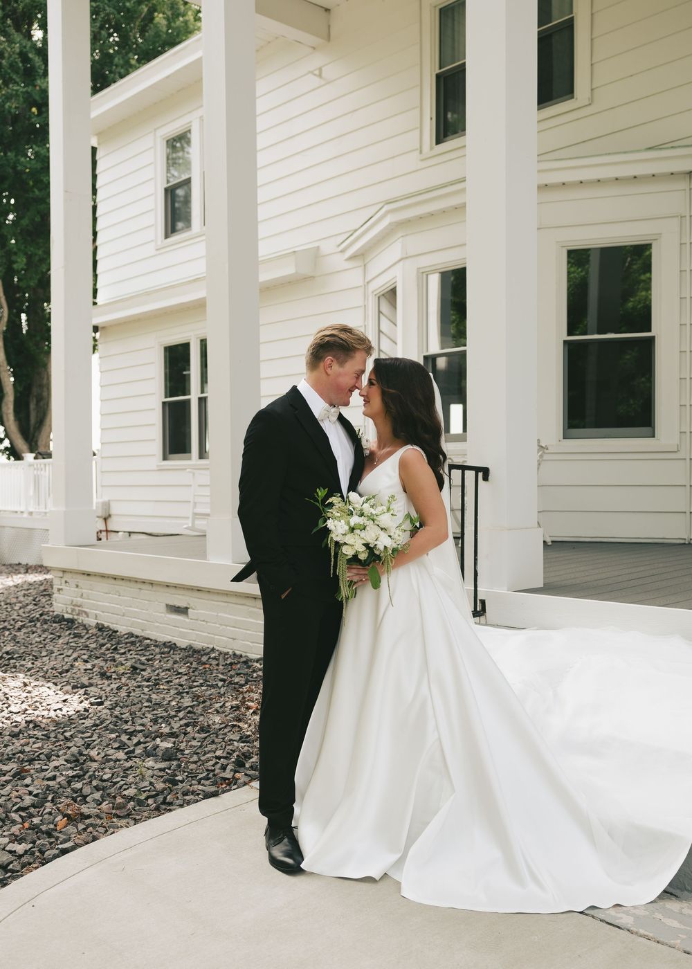 Bride and groom share a romantic moment outside a white colonial wedding venue, bride holding a white floral bouquet.