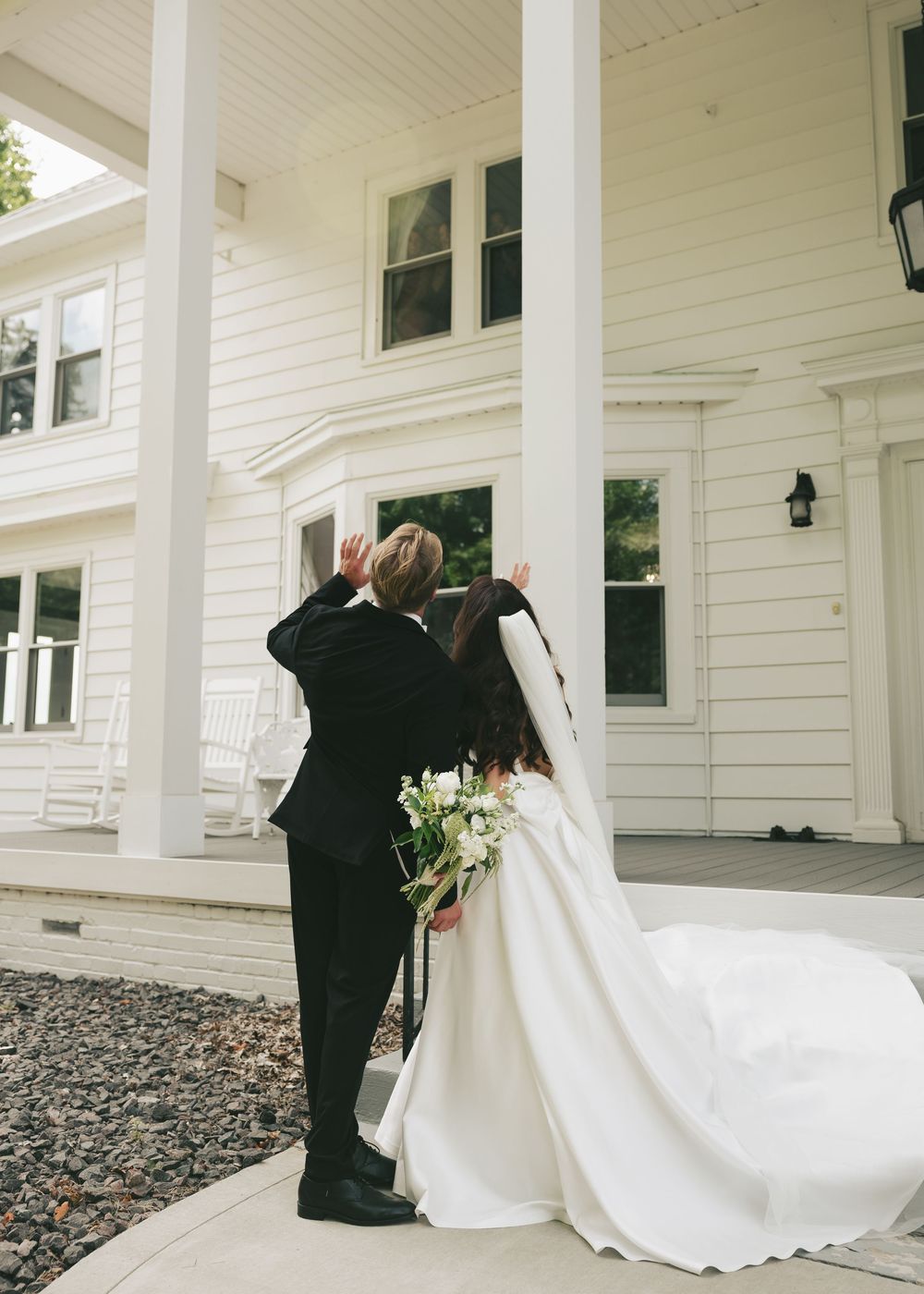 A bride and groom share a romantic moment facing a white colonial home, her elegant gown trailing behind them.