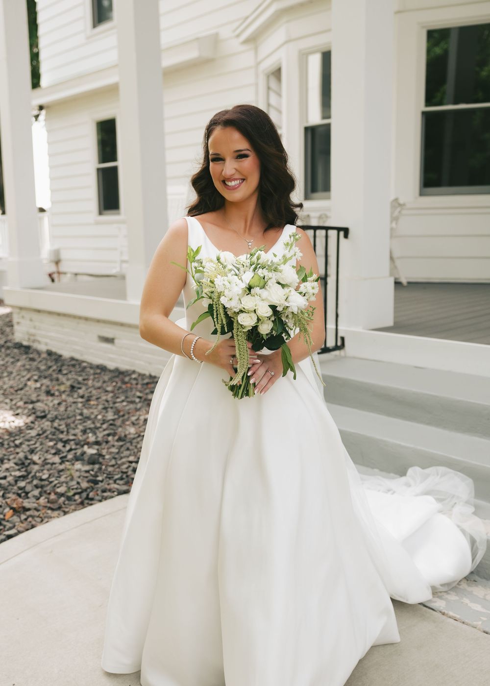Smiling bride in elegant white ball gown holding white floral bouquet, standing in front of a classic white mansion.
