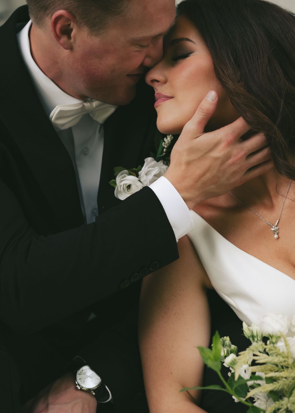 Romantic bride and groom sharing an intimate moment, groom in black tuxedo with white boutonniere gently holding brides face.