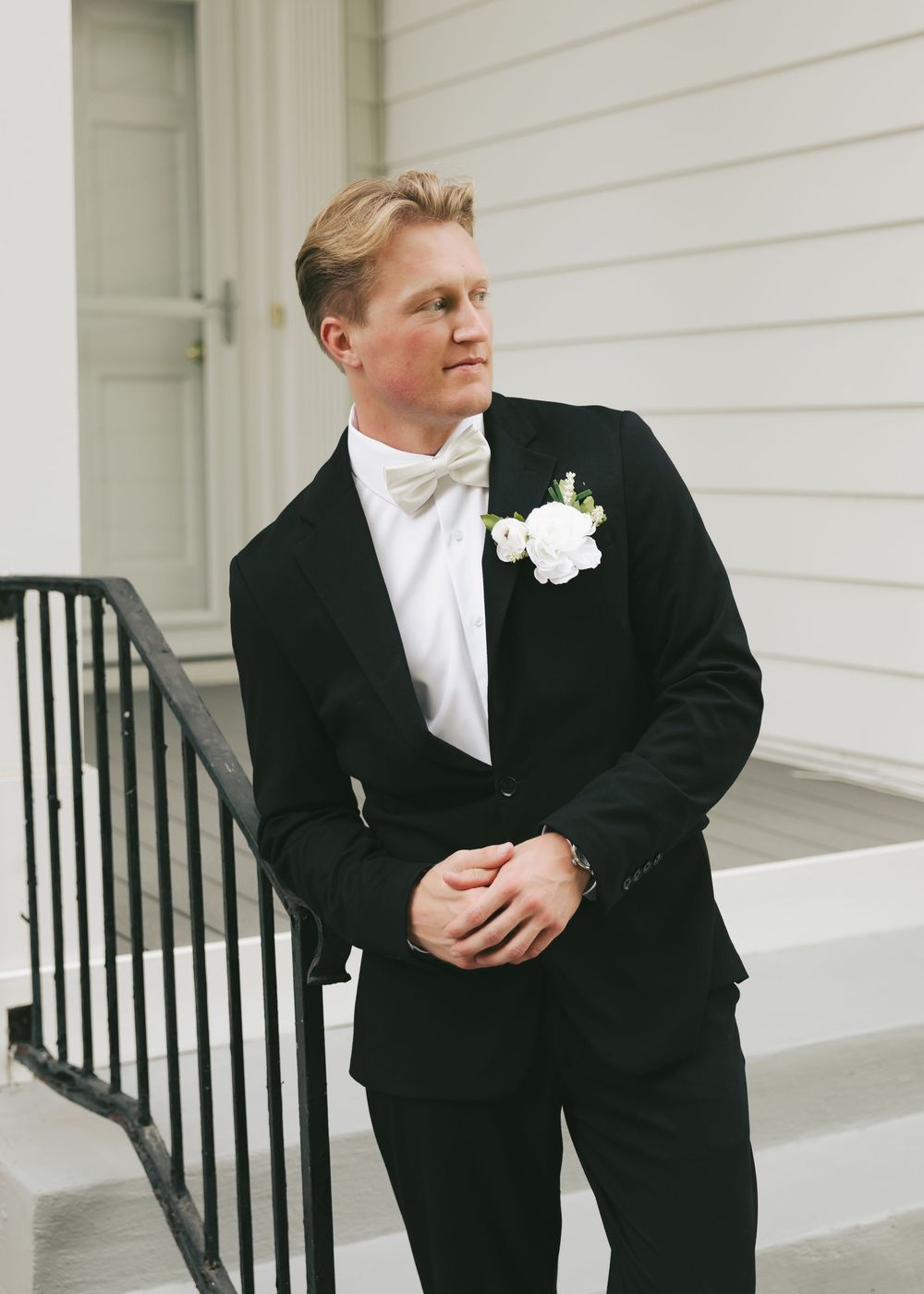 A groom in a black tuxedo with white bow tie and floral boutonniere stands on white steps gazing away.