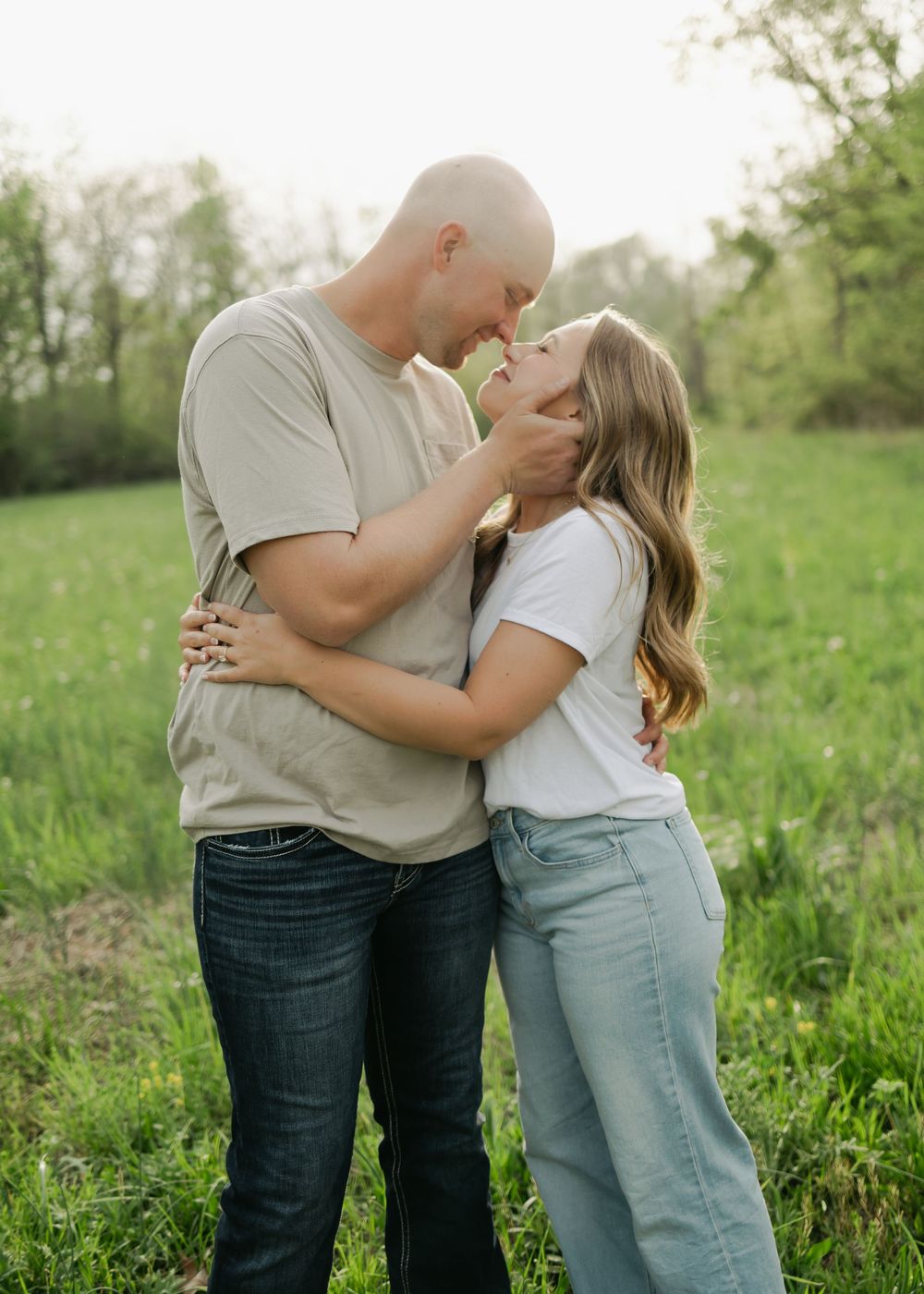 Engaged couple embracing and touching foreheads tenderly in a lush green meadow at golden hour.