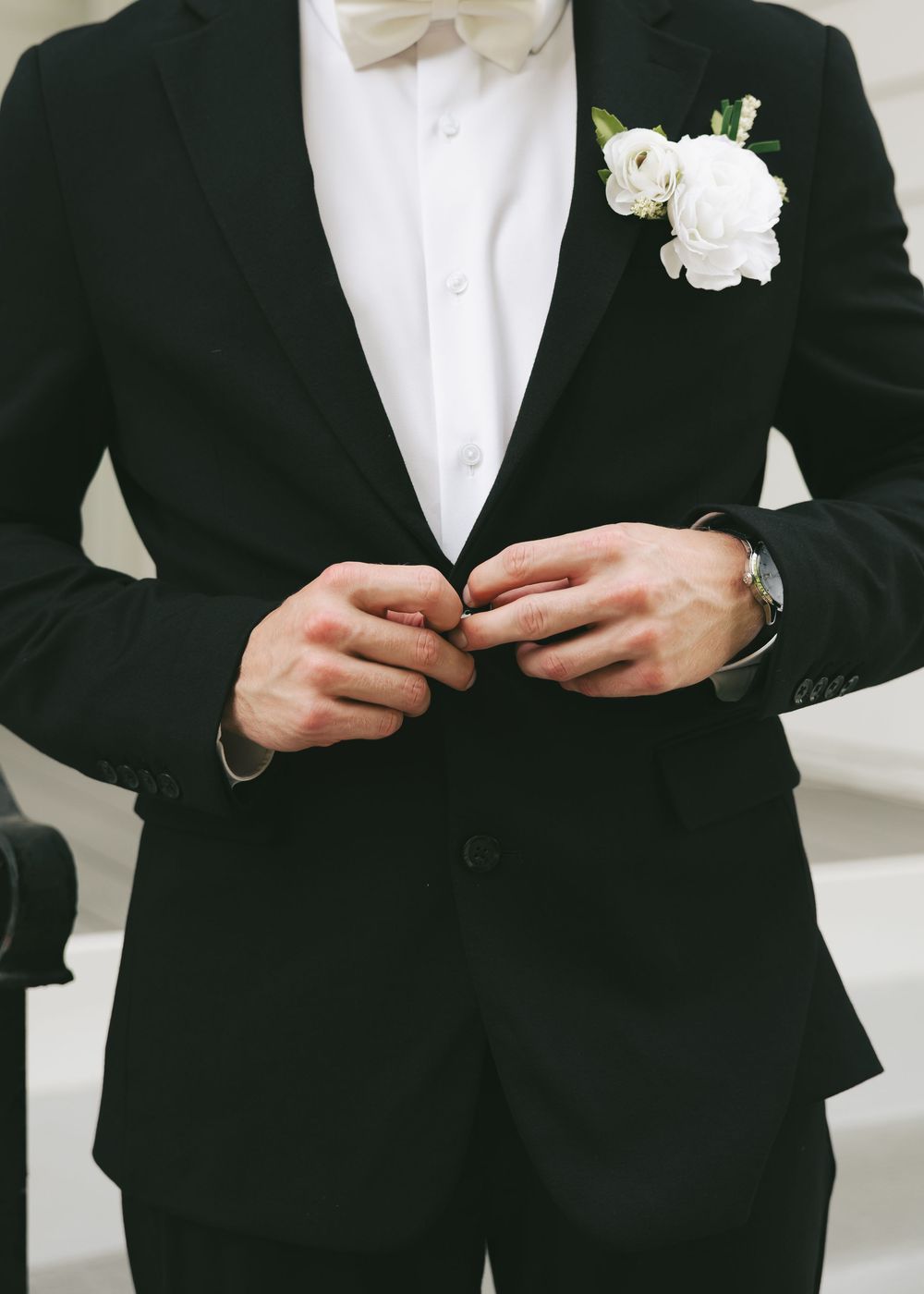 Groom in elegant black tuxedo with white bow tie and floral boutonniere buttoning his jacket before wedding ceremony.