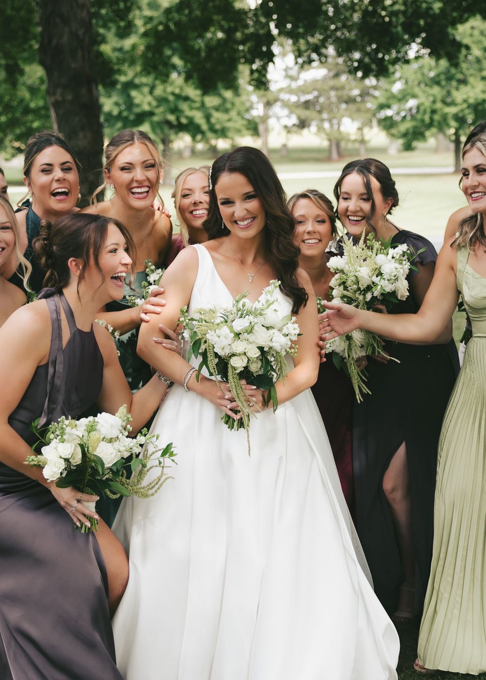 Bride in white gown holds white bouquet surrounded by laughing bridesmaids in mixed green and gray dresses outdoors.