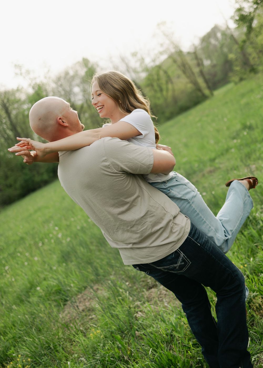 Man lifting and spinning his laughing fiancee in a green meadow during a fun outdoor engagement session.