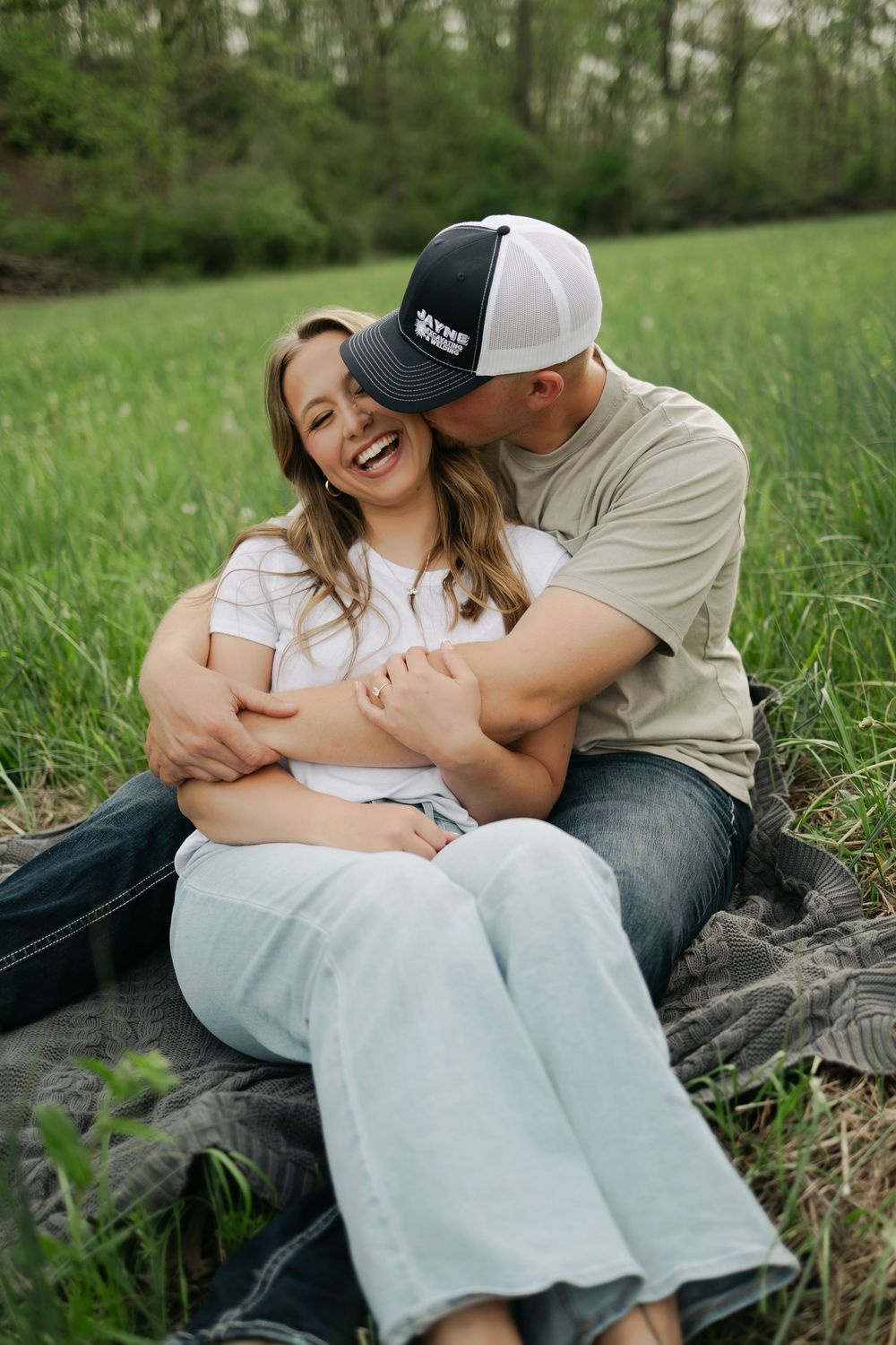 Couple sitting on a blanket in tall grass as man in trucker hat kisses smiling womans cheek.