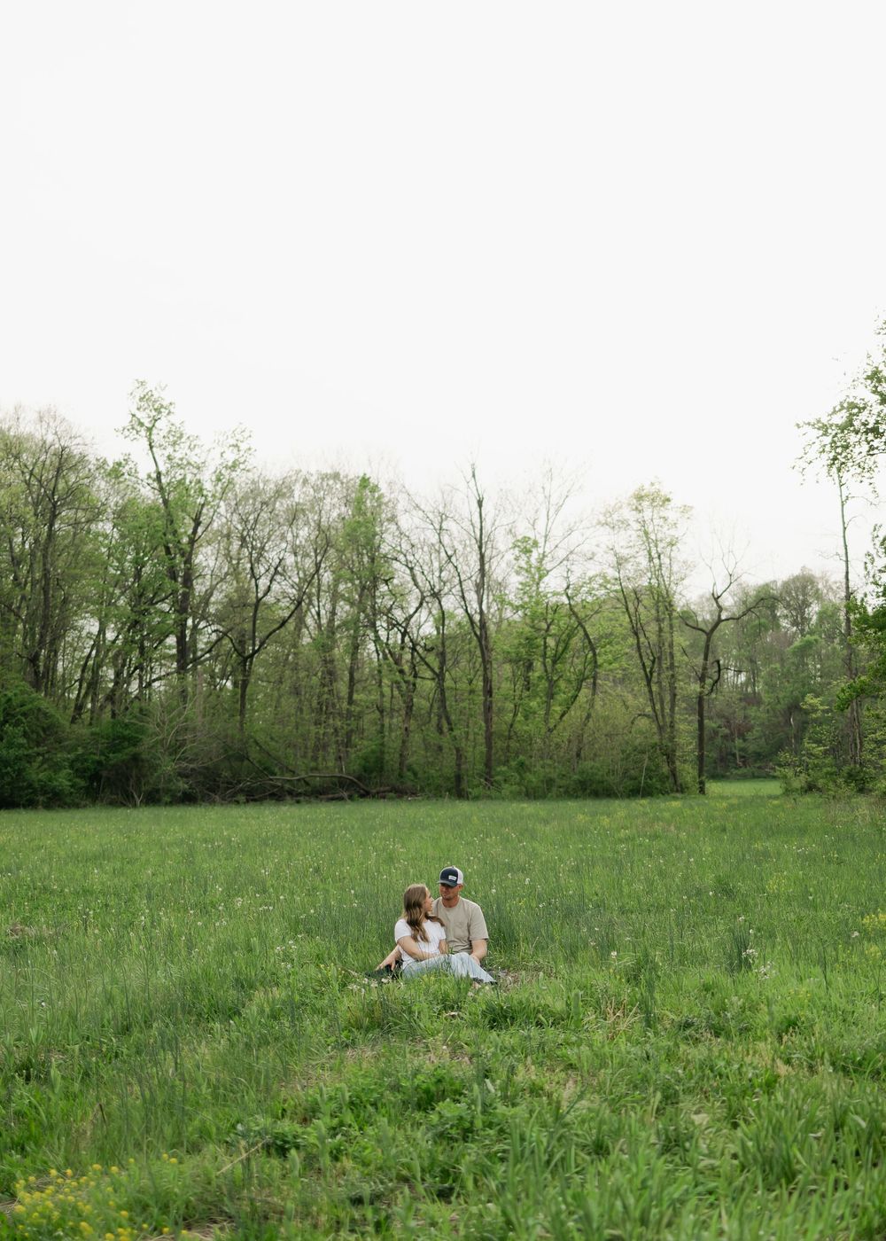 Engaged couple sitting together in a vast green meadow surrounded by trees during spring engagement photos.