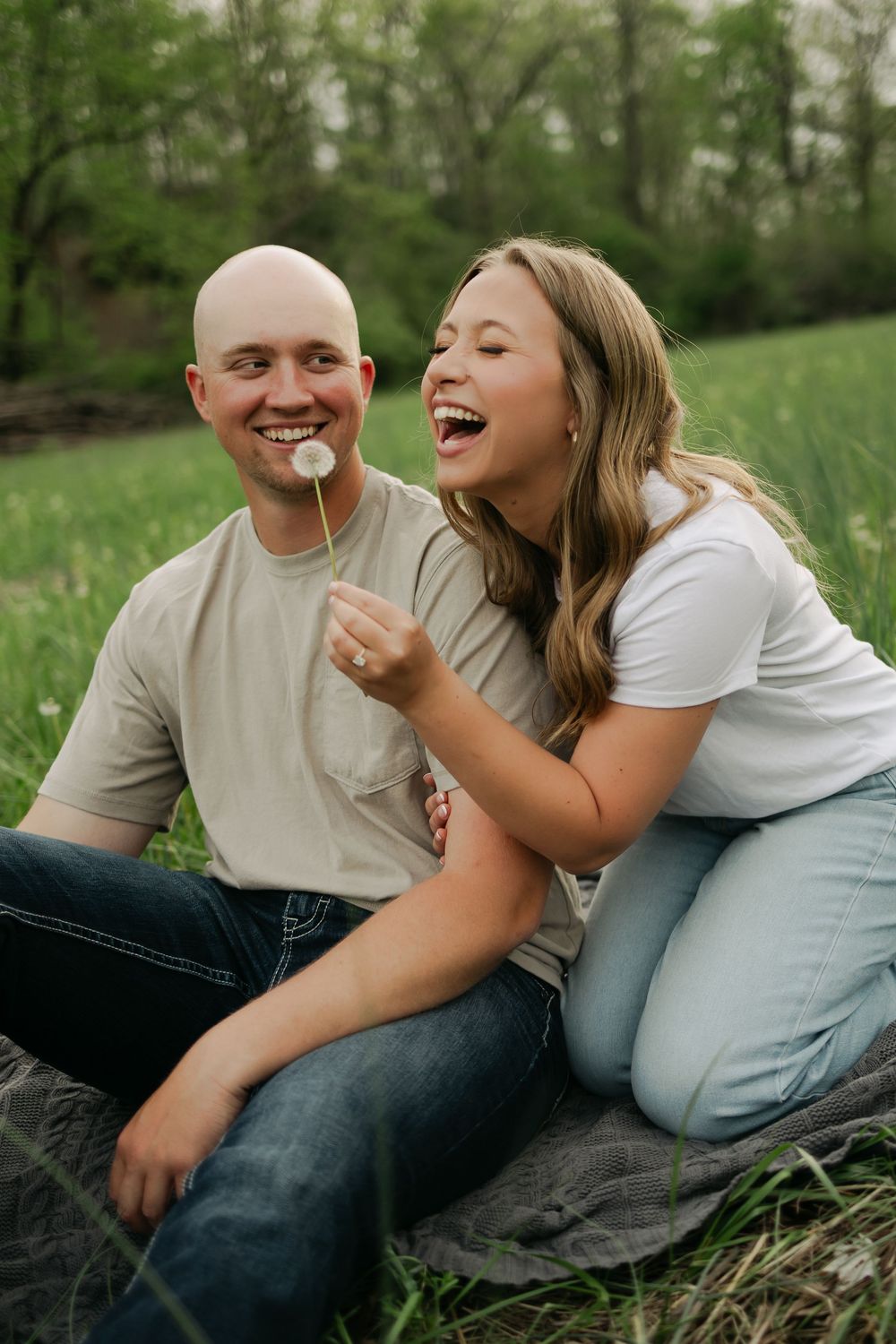 Laughing couple sitting in a field as woman playfully holds a dandelion up to her fiances face.