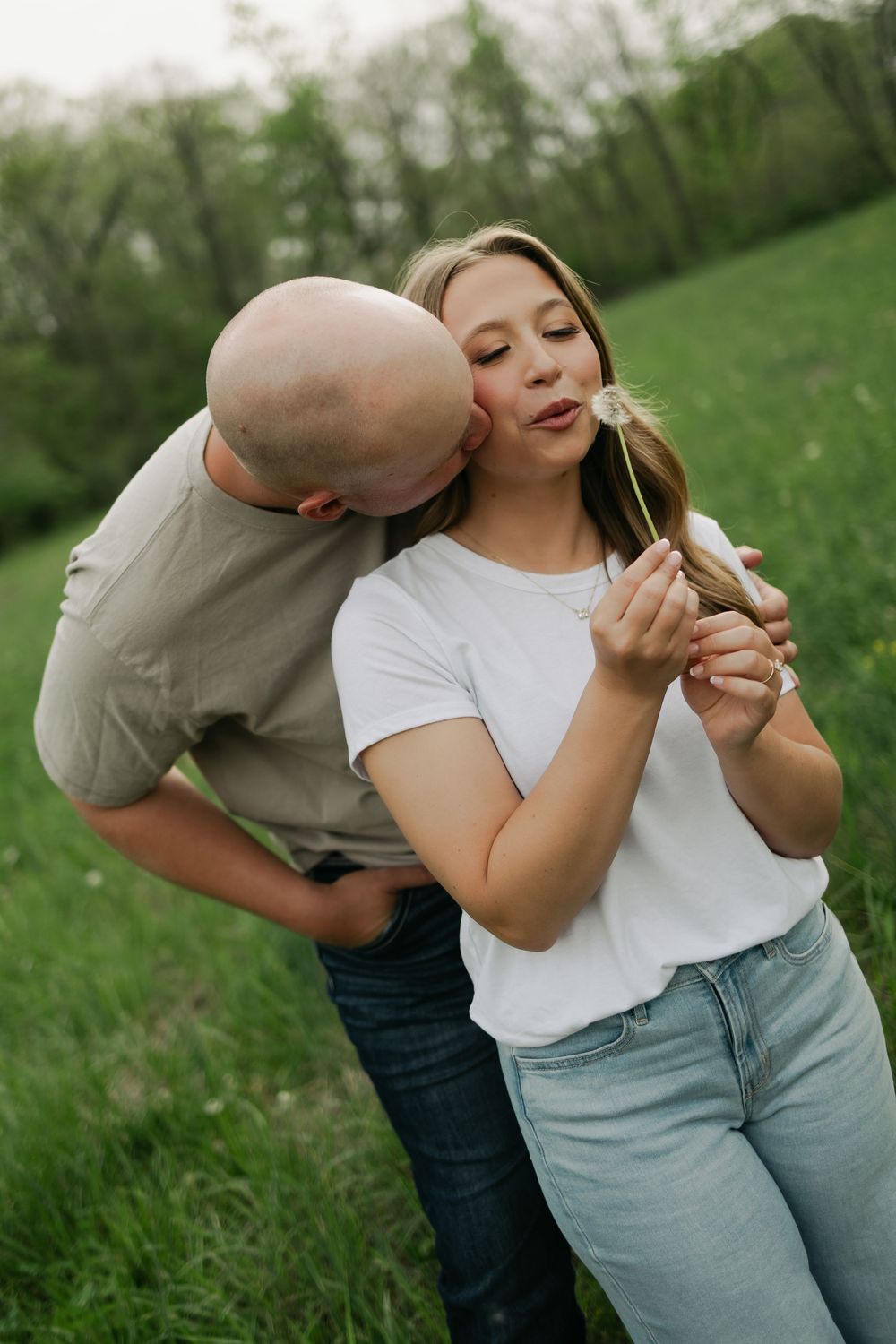 Woman blows a dandelion wish while her partner kisses her cheek in a lush green outdoor setting.