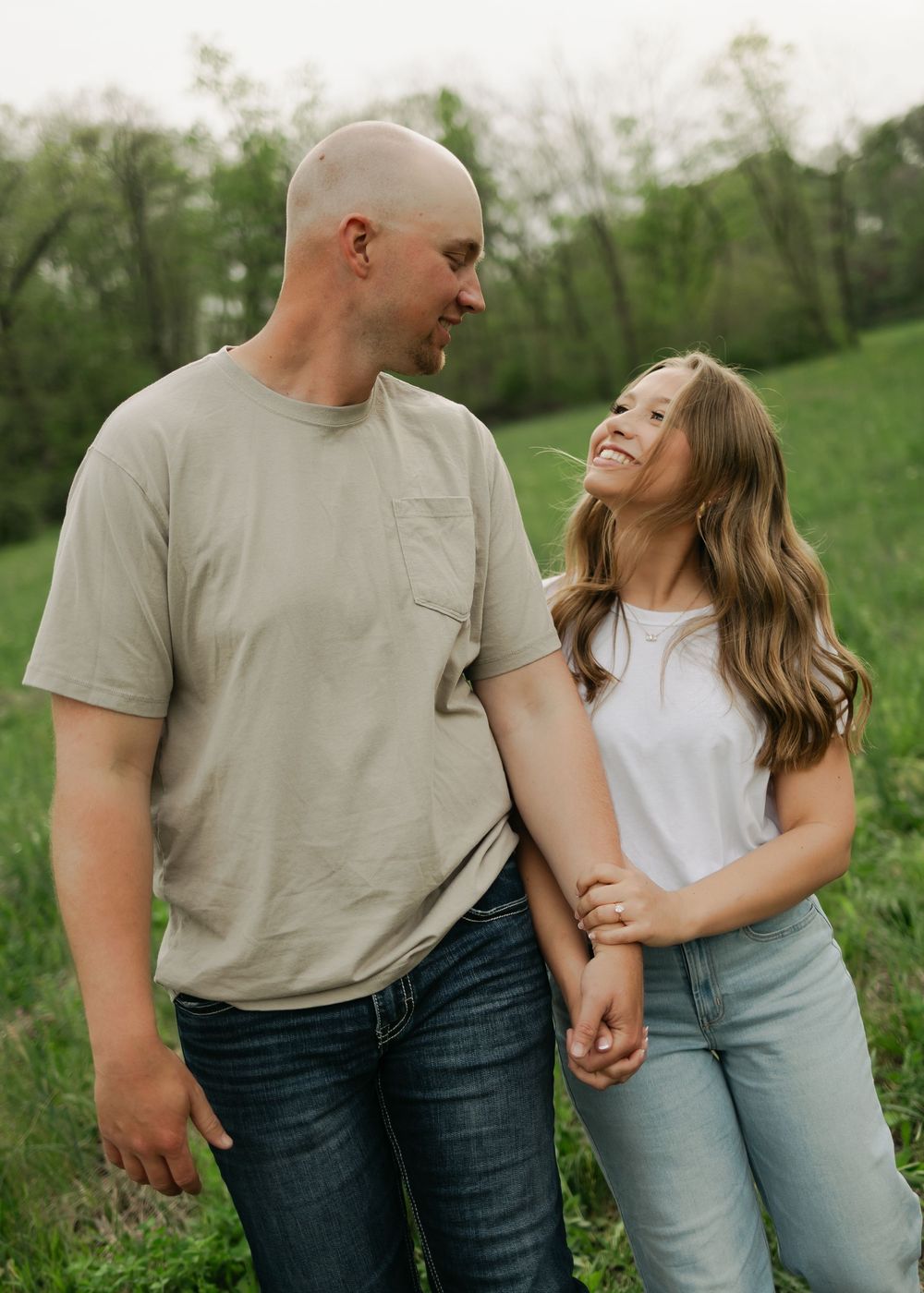 Couple holding hands and smiling at each other in a green field during an outdoor engagement photo session.
