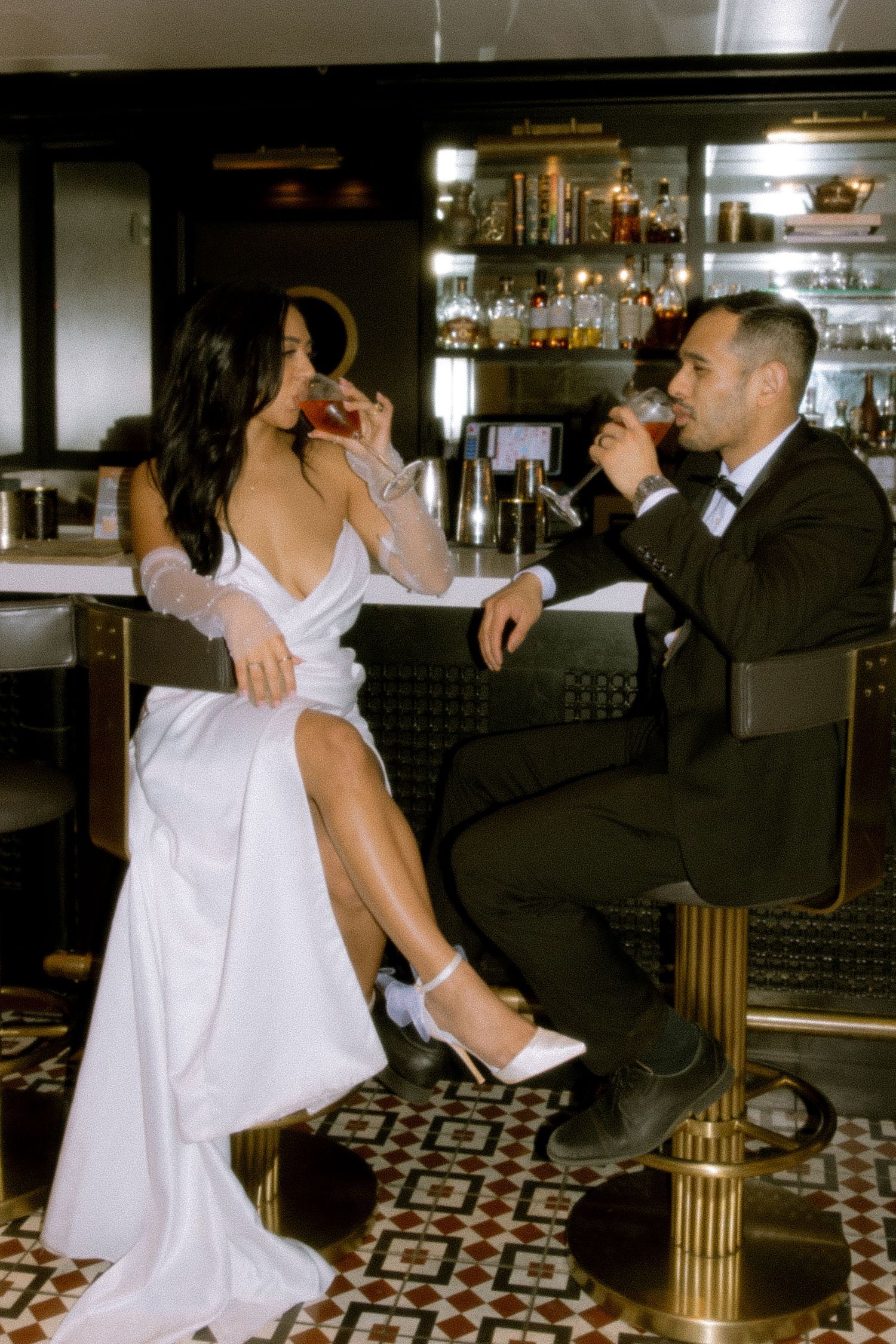 Bride and groom sipping cocktails at a stylish bar, bride in white gown with slit, groom in black tuxedo.