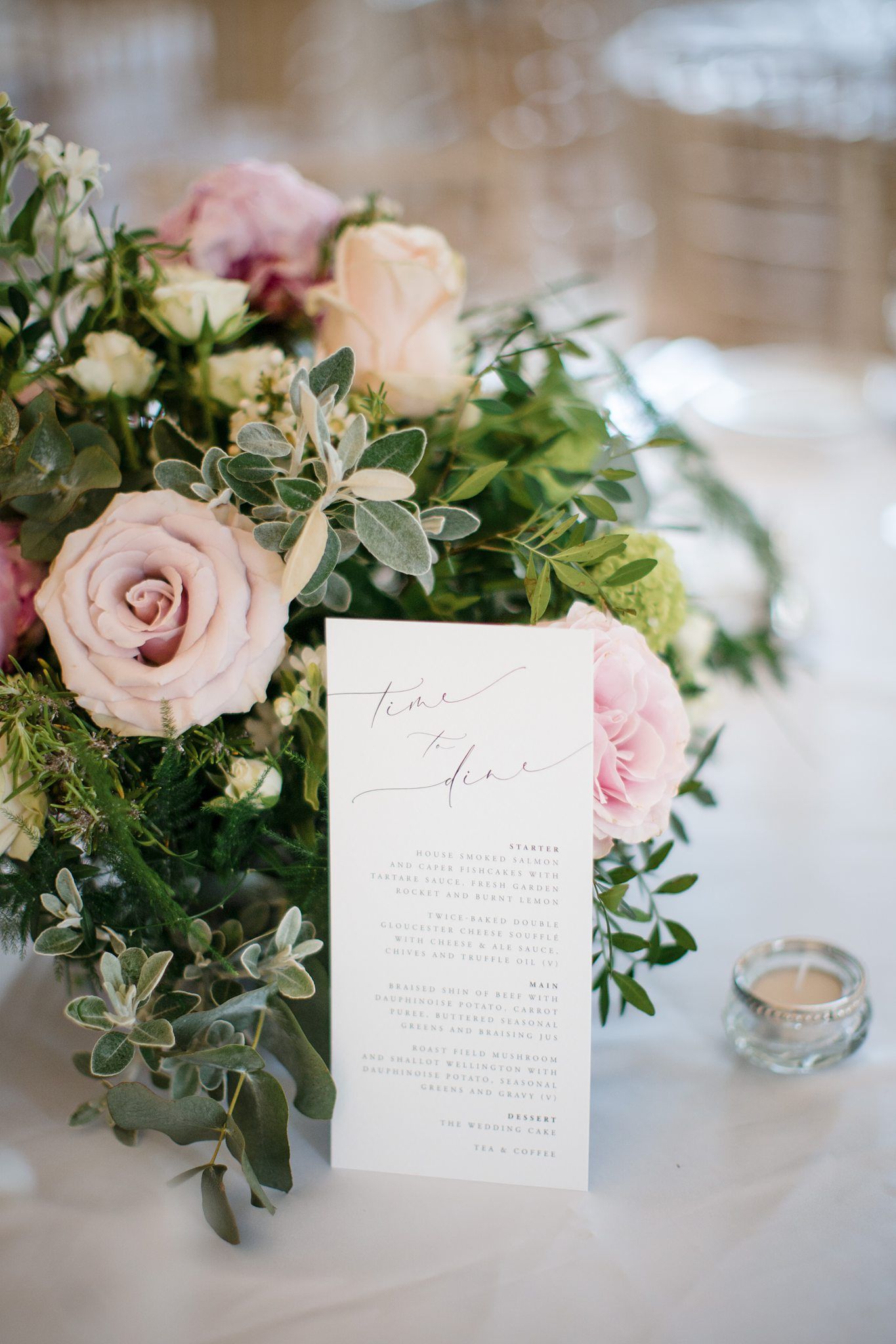 Elegant wedding reception table with pink roses, greenery centerpiece, and a calligraphy menu card beside a tealight candle.