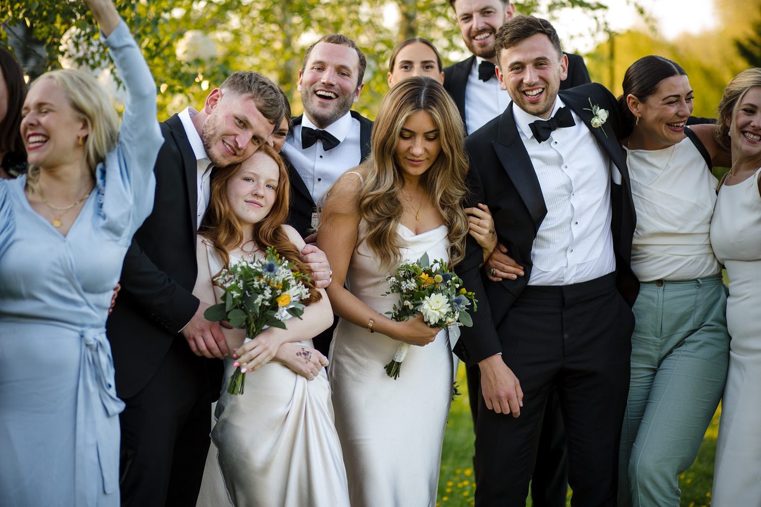 Bride and groom celebrate with wedding party in formal attire outdoors, smiling and cheering with bouquet.