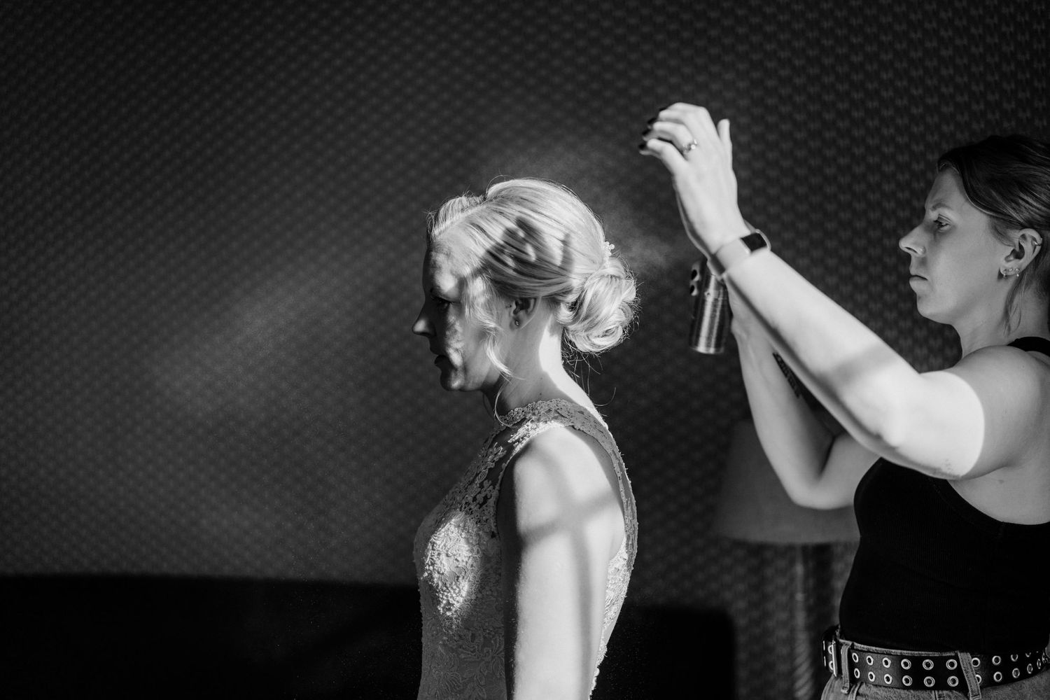 Black and white photo of a hairstylist spraying hairspray on a blonde womans updo hairstyle backstage.