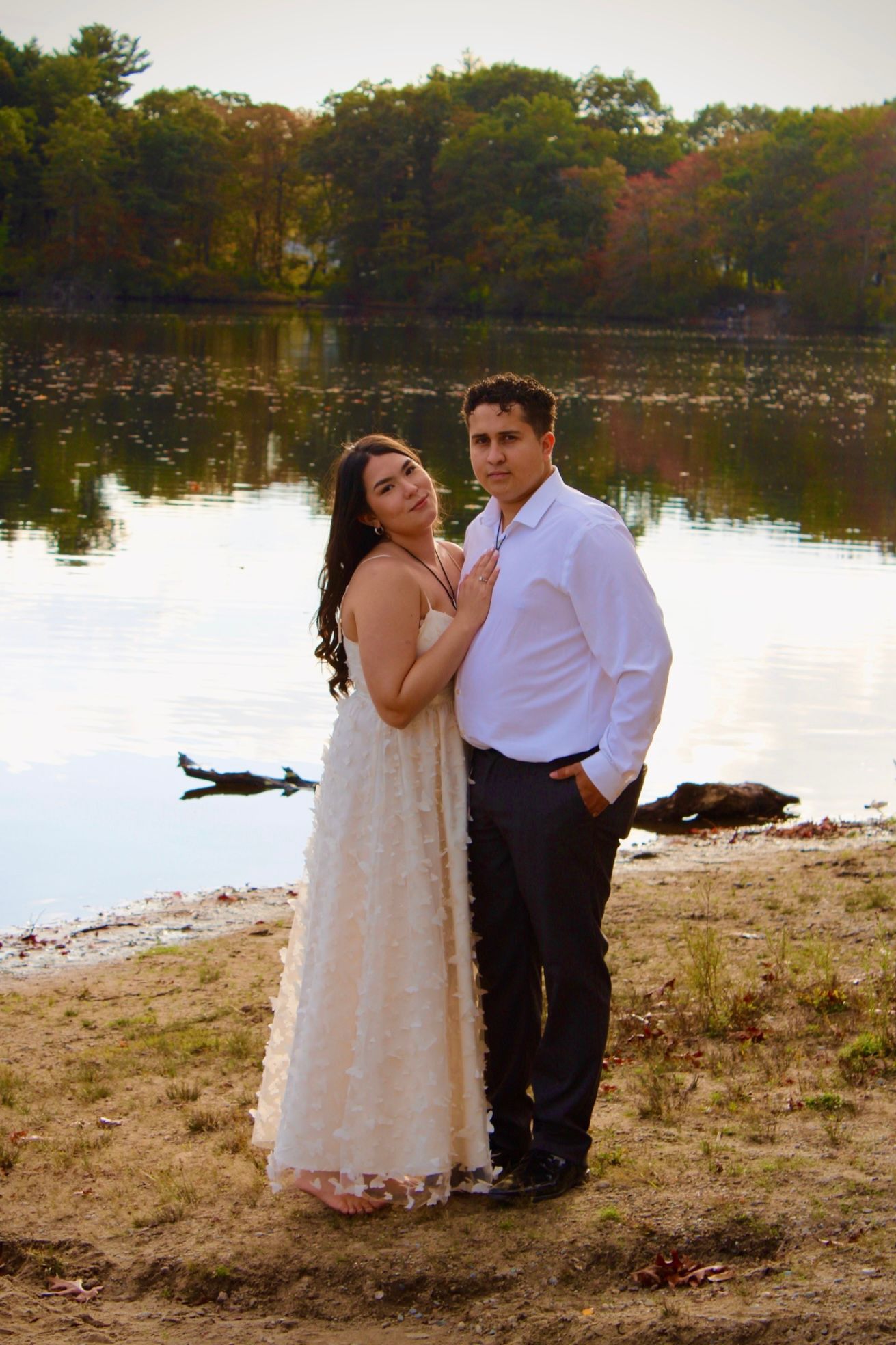 A romantic couple in formal attire shares tender moments by a serene lake during autumn sunset for their engagement photoshoot.
