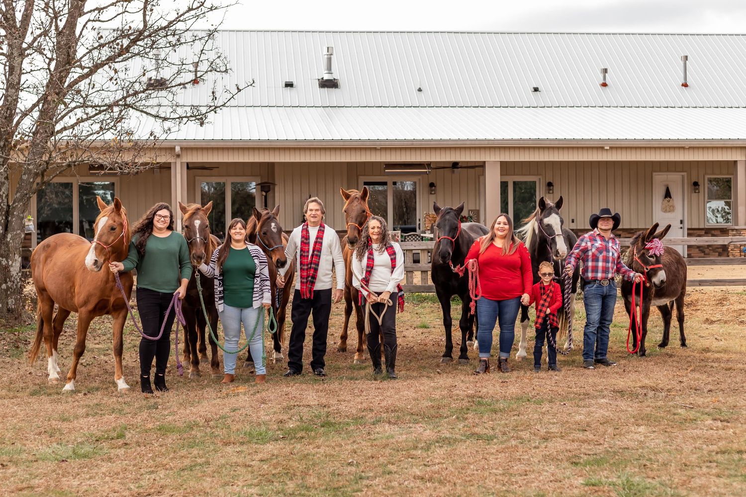 Large group standing with horses outside a rustic barn building.