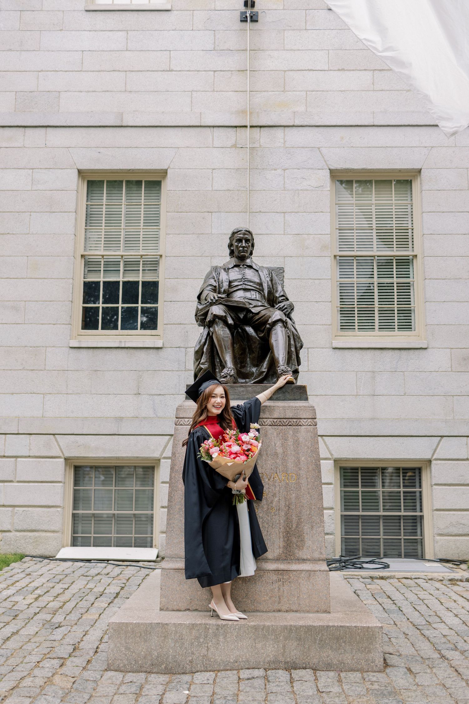 Graduate in cap and gown holding flowers poses before the John Harvard statue at Harvard University.