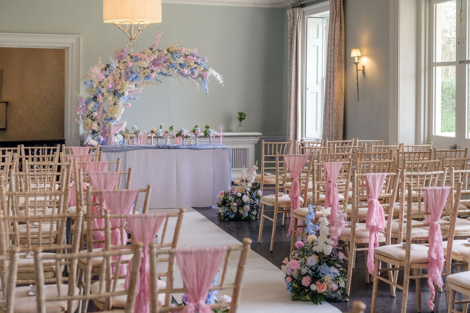 Elegant wedding ceremony room with gold chiavari chairs, pink sashes, floral aisle decor, and a stunning circular flower arch backdrop.
