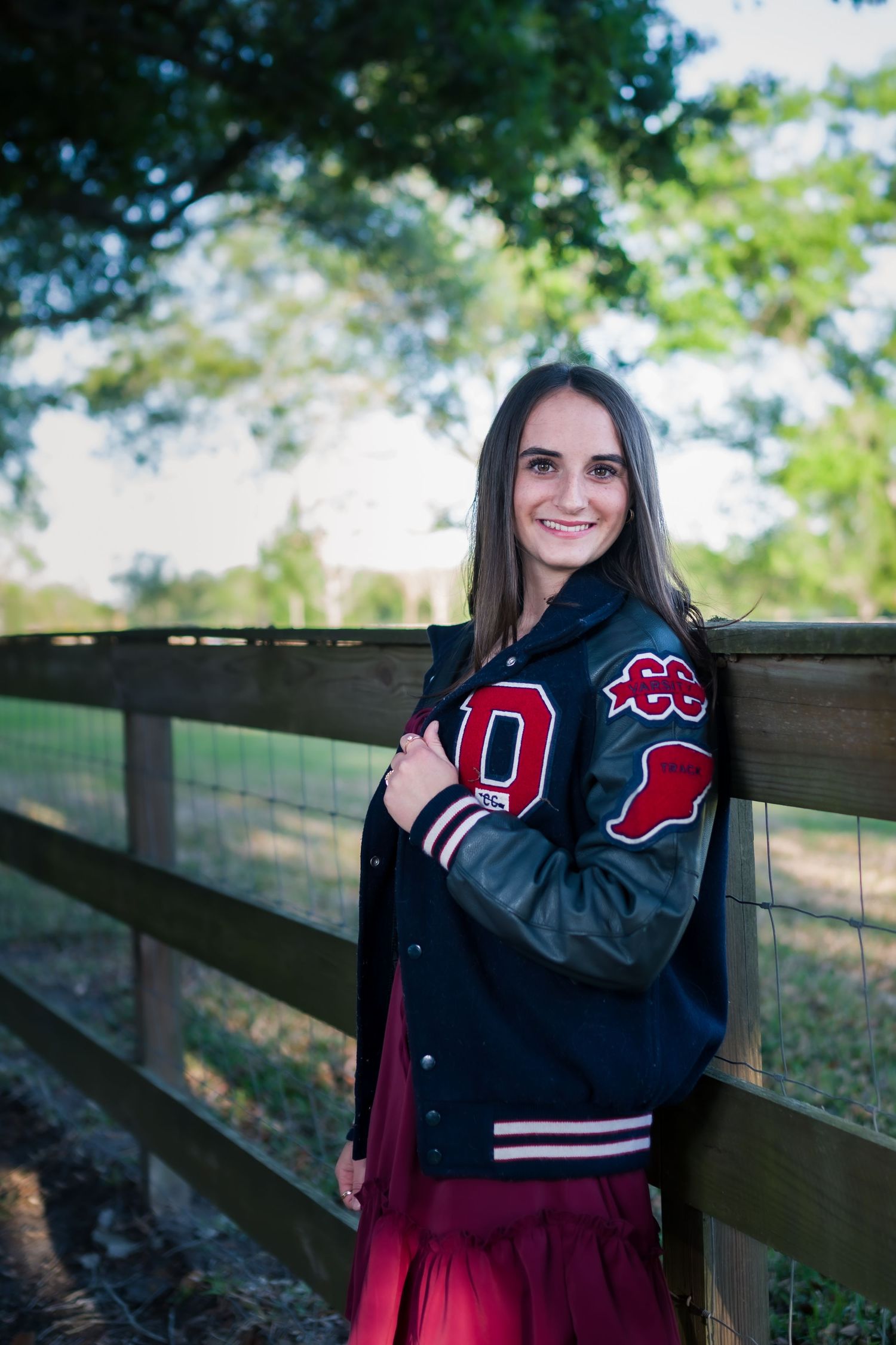 Young woman in varsity jacket and red skirt smiling by wooden fence outdoors.